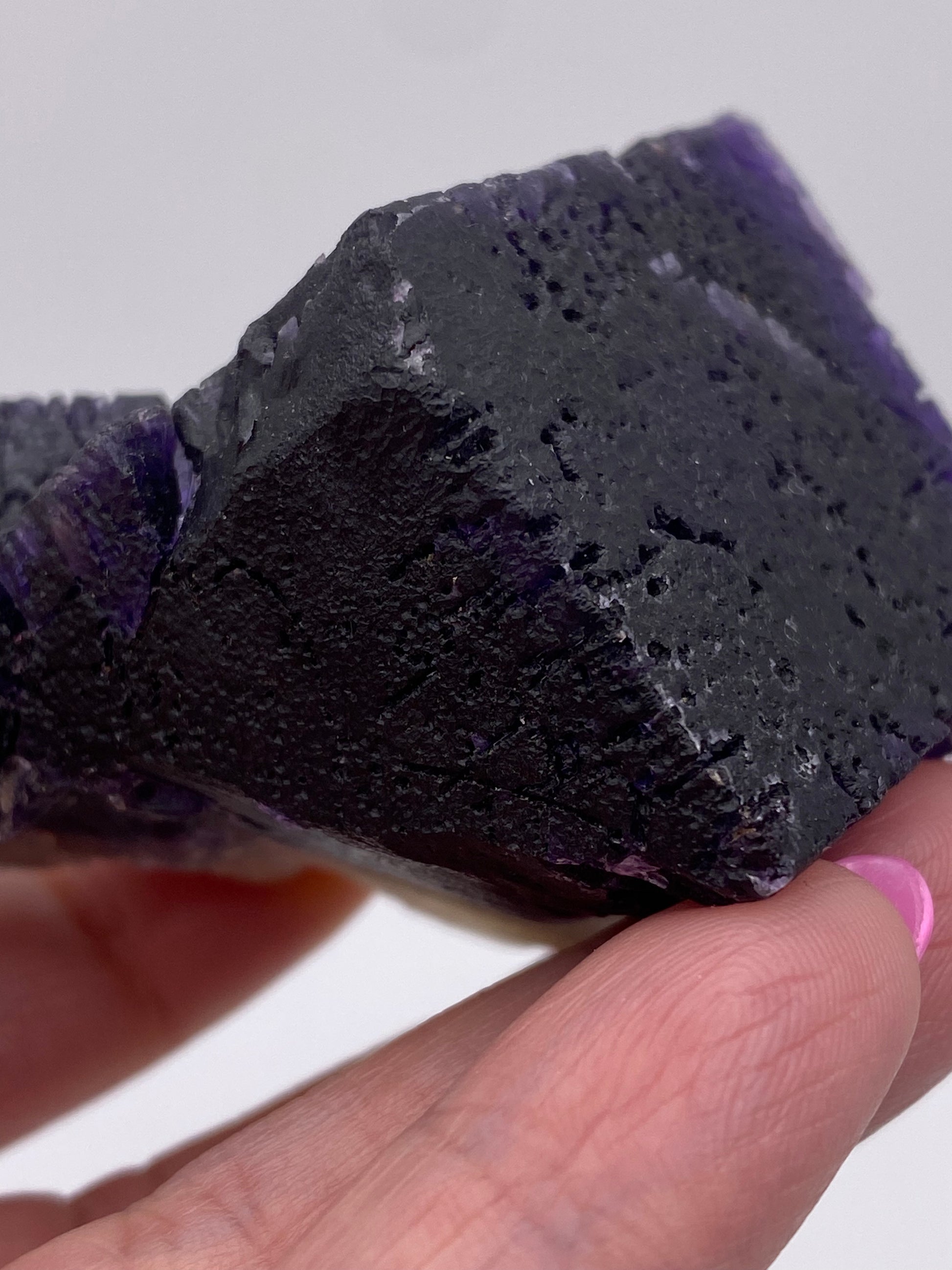 A hand holds a rough, rectangular piece of dark-colored mineral rock with jagged edges and a slightly porous surface. This specimen is Fluorite from the Hill-Ledford Mine in Cave-In-Rock, Hardin County, Illinois, USA. The crystal exhibits patches of deep purple hues typical of fluorite. The fingernails are painted pink, and the background is plain white. This exquisite piece is brought to you by The Crystalary.