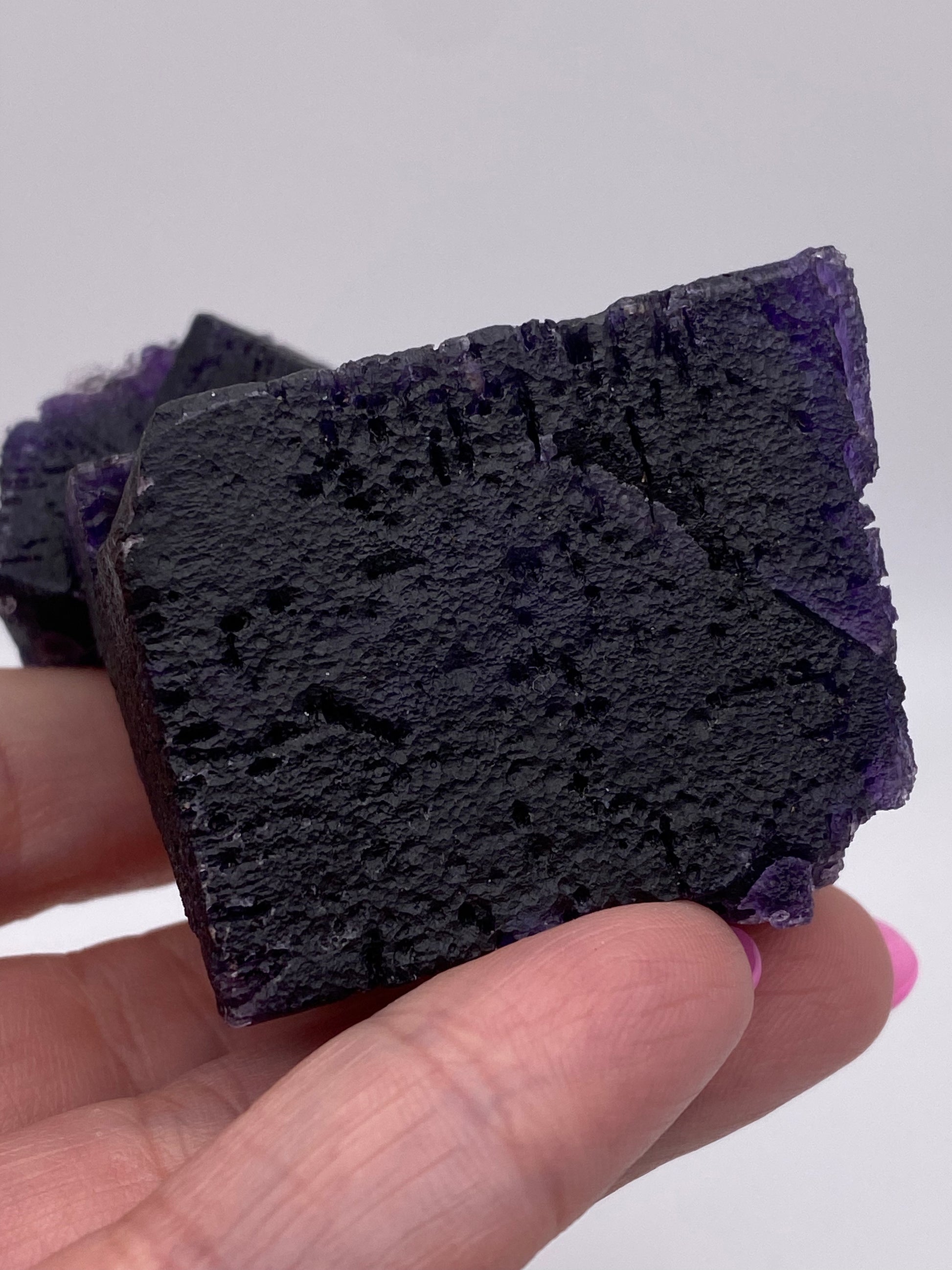 A person holds The Crystalary's Fluorite from the Hill-Ledford Mine in Cave-In-Rock, Hardin County, Illinois, USA against a plain, off-white background. The dark purple mineral specimen has a rough and irregular cubic shape with shades of purple visible across its textured surface. The person's fingers are partially seen with a hint of pink nail polish.