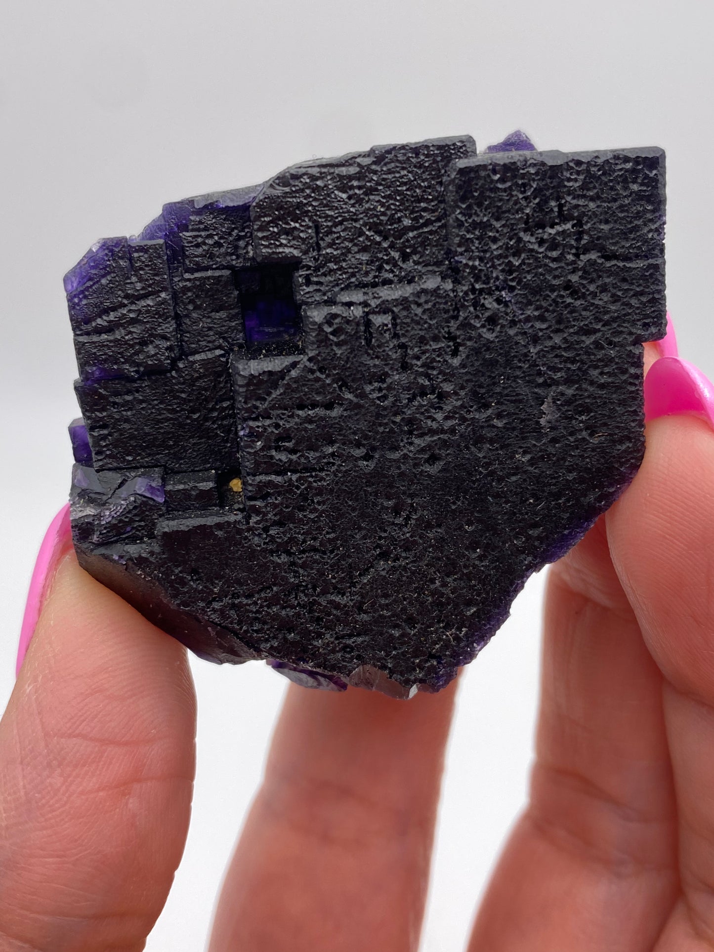 A Fluorite from The Crystalary, specifically sourced from the Hill-Ledford Mine in the Cave-In-Rock Mining Sub-District of Hardin County, Illinois, USA, is being held between the thumb and index finger of a person with pink nail polish. The mineral appears black with a slight purple hue and features an uneven and jagged surface. The background is plain white.