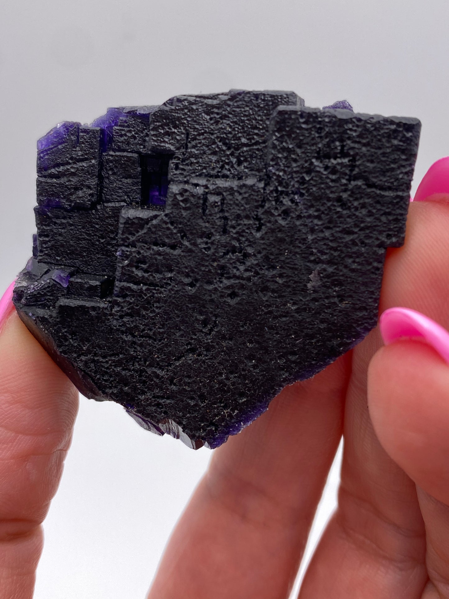 A close-up photo highlights two hands with pink-painted nails holding a rough, black crystal with hints of deep purple. The background is white and out of focus, emphasizing the dark crystal's texture and uneven edges. This stunning Fluorite from The Crystalary comes from the Hill-Ledford Mine in Hardin County's renowned Cave-In-Rock Mining Sub-District, Illinois, USA.