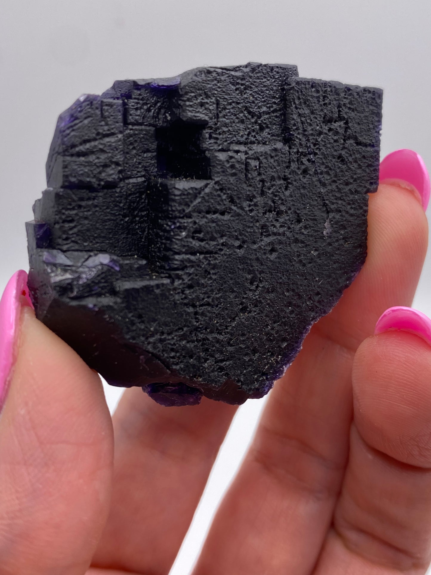 A close-up of a hand holding a rough, dark purple Fluorite specimen from The Crystalary, sourced from the Hill-Ledford Mine in the Cave-In-Rock Mining Sub-District of Hardin County, Illinois. The mineral boasts a textured surface with visible layers and edges, creating a striking contrast against the hand’s bright pink nail polish. The background is blurred and neutral.