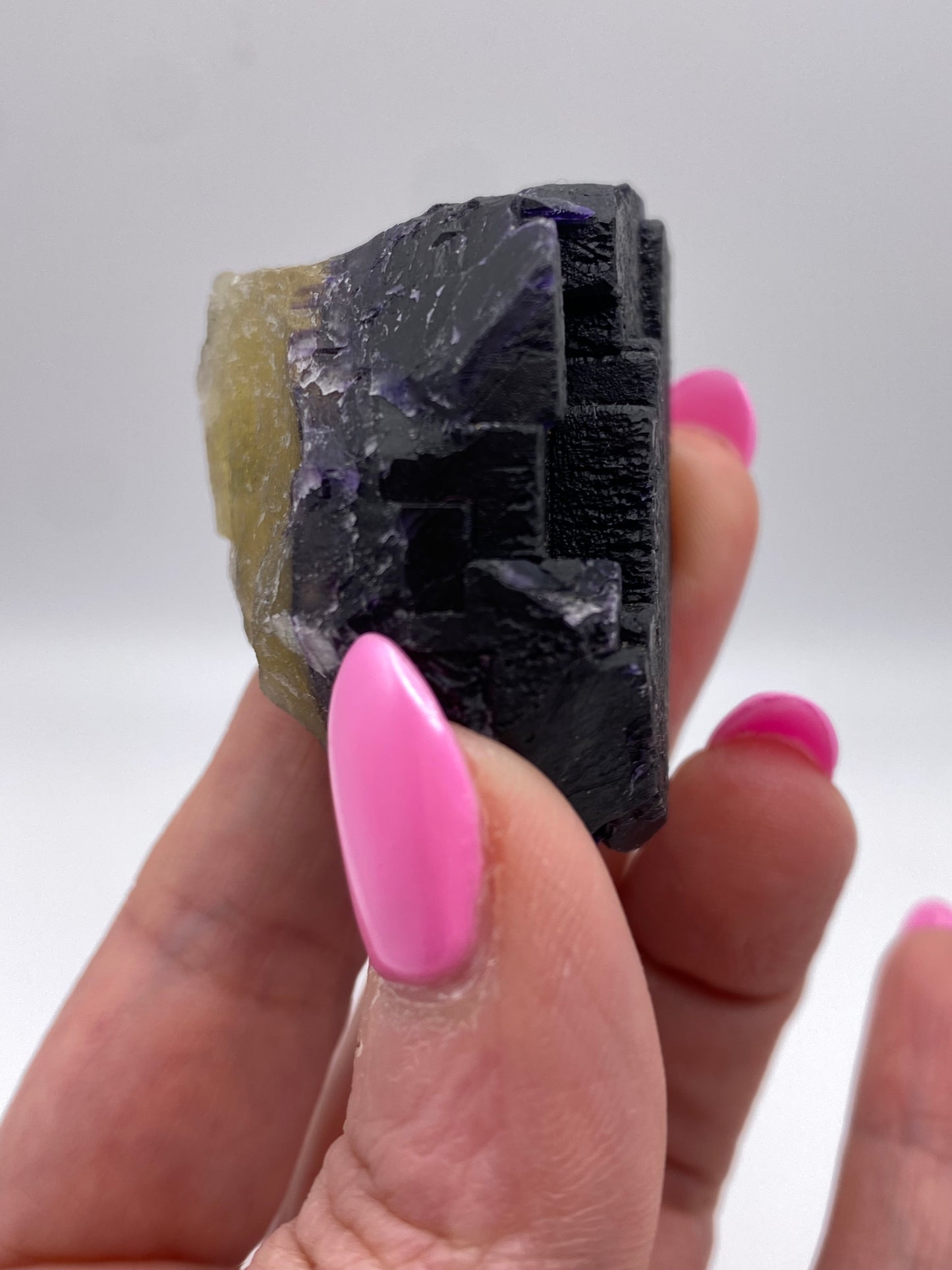 A hand with bright pink nails holds a small, angular Fluorite specimen from The Crystalary's Hill-Ledford Mine in the Cave-In-Rock Mining Sub-District, Hardin County, Illinois. The mineral boasts a dark, shiny surface adorned with visible geometric formations and lighter, semi-transparent patches. A softly blurred background enhances the focus on the exquisite mineral.