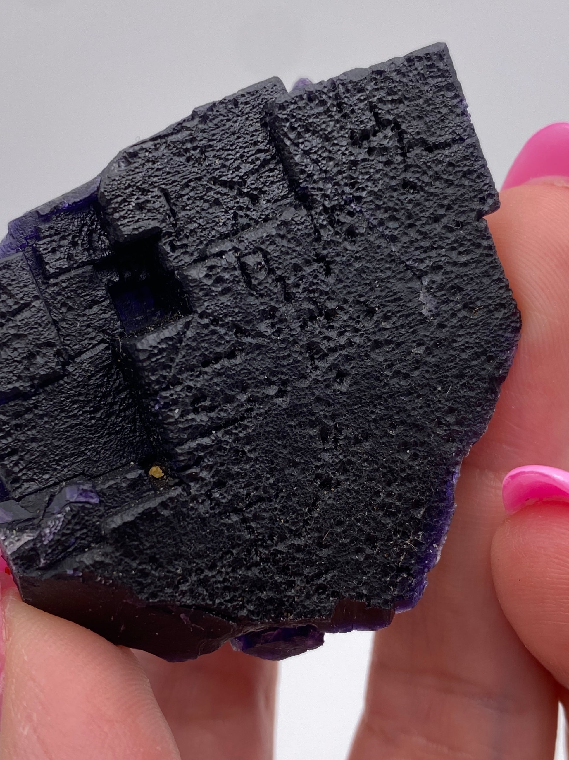 A close-up image of a black, rough-textured crystal from The Crystalary's Fluorite collection, specifically sourced from the Hill-Ledford Mine in the Cave-In-Rock Mining Sub-District of Hardin County, Illinois. The geometric, blocky crystal is held between two fingers with pink-painted nails and set against a plain white background.