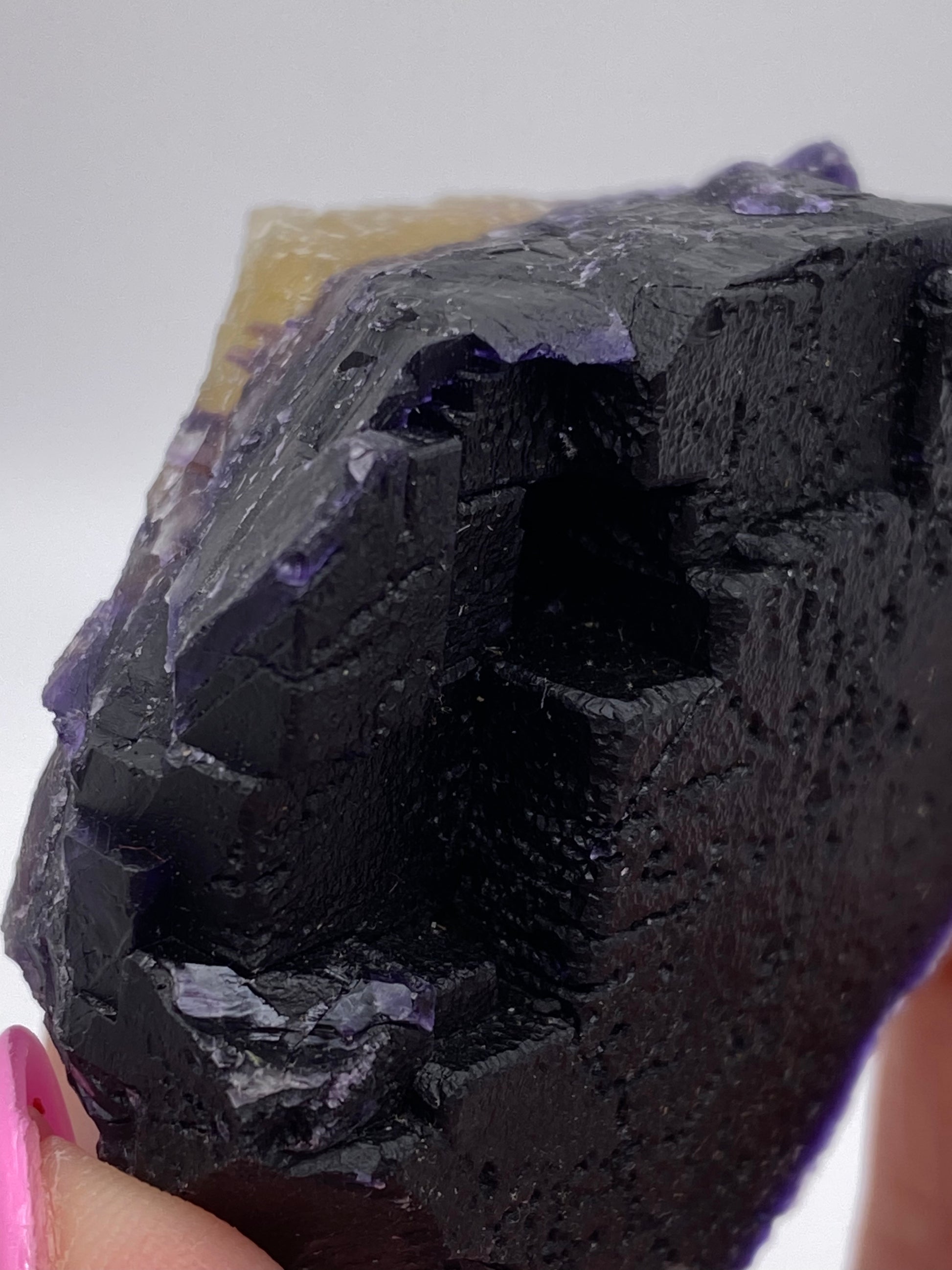 Close-up of a dark, jagged fluorite from The Crystalary's Hill-Ledford Mine collection, with a textured surface, held between fingers with pink-painted nails. The mineral boasts deep black and purple hues, light reflecting off its uneven edges. The background is blurred and minimal, highlighting the exquisite find from Hardin County’s Cave-In-Rock Mining Sub-District in Illinois, USA.