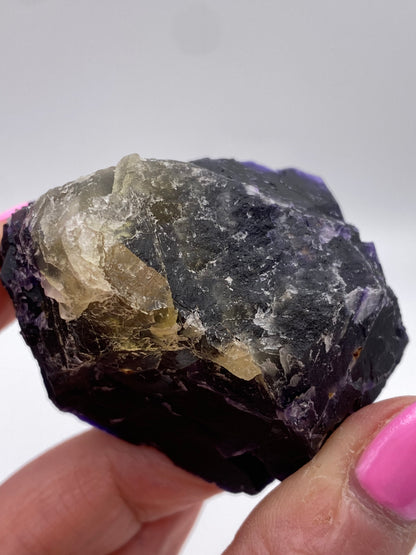 A hand with pink nail polish is holding a rugged, dark-colored Fluorite from the Hill-Ledford Mine in the Cave-In-Rock Mining Sub-District, Hardin County, Illinois. The stone, branded by The Crystalary, features whitish translucent sections and purple hues along its jagged and uneven edges. The background is plain and out of focus, drawing attention to this unique mineral specimen.