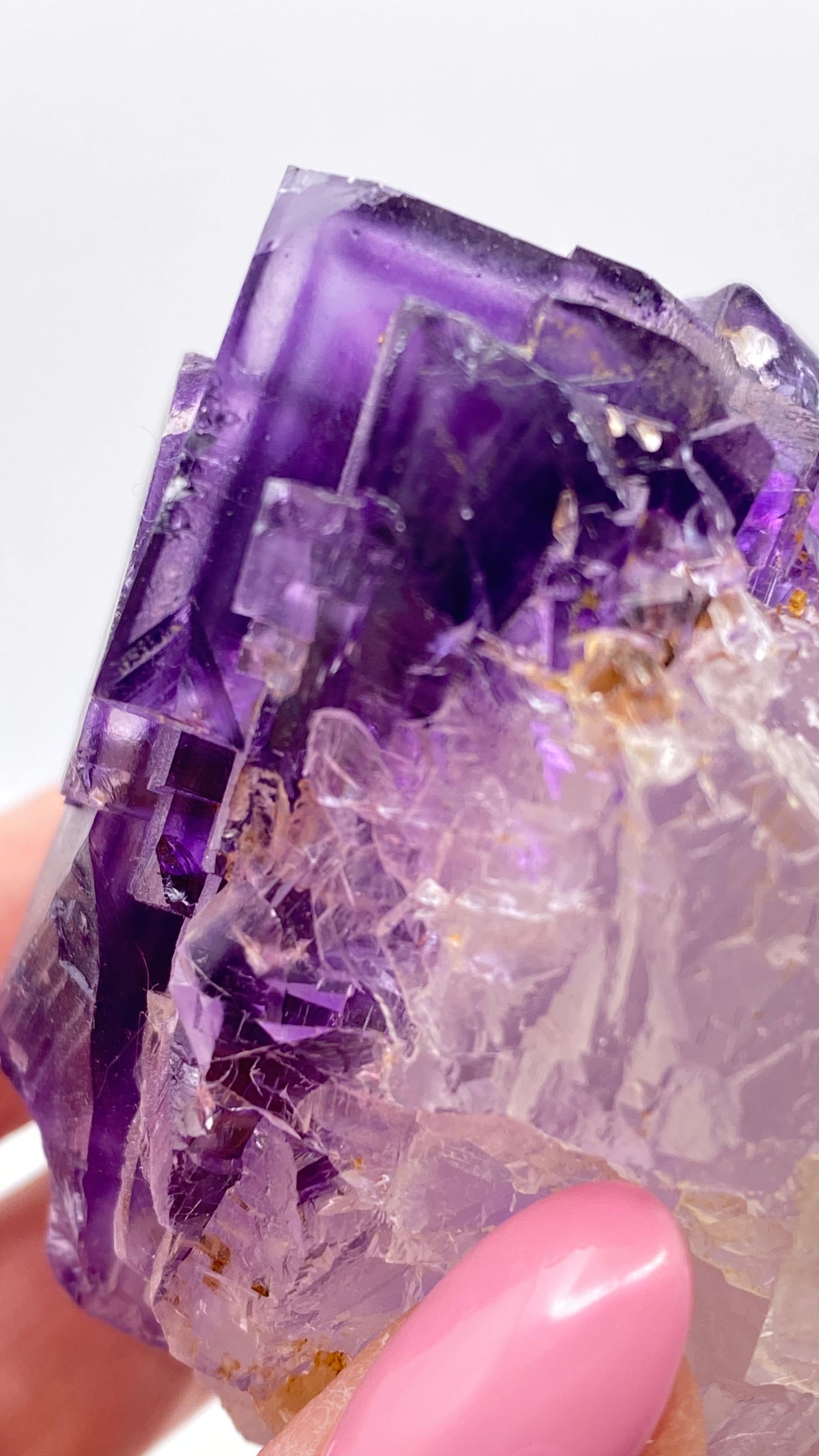 A close-up photo showcases a hand holding a stunning Fluorite crystal from The Crystalary, featuring purple and translucent facets. The geometric shapes and intricate textures of the crystal from Lead Hill, Cave-In-Rock, Hardin County, Illinois, USA are accentuated by the person's light pink painted fingernail.