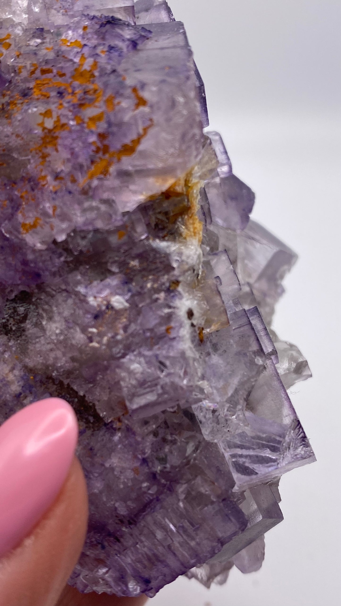 A close-up photograph of a purple crystalline mineral, likely Fluorite- Lead Hill from Cave-In-Rock Mining in Hardin County by The Crystalary, held by a person with a pink manicured thumb visible in the bottom left. The mineral has a rough texture with orange and white inclusions. The background is a plain, soft white.