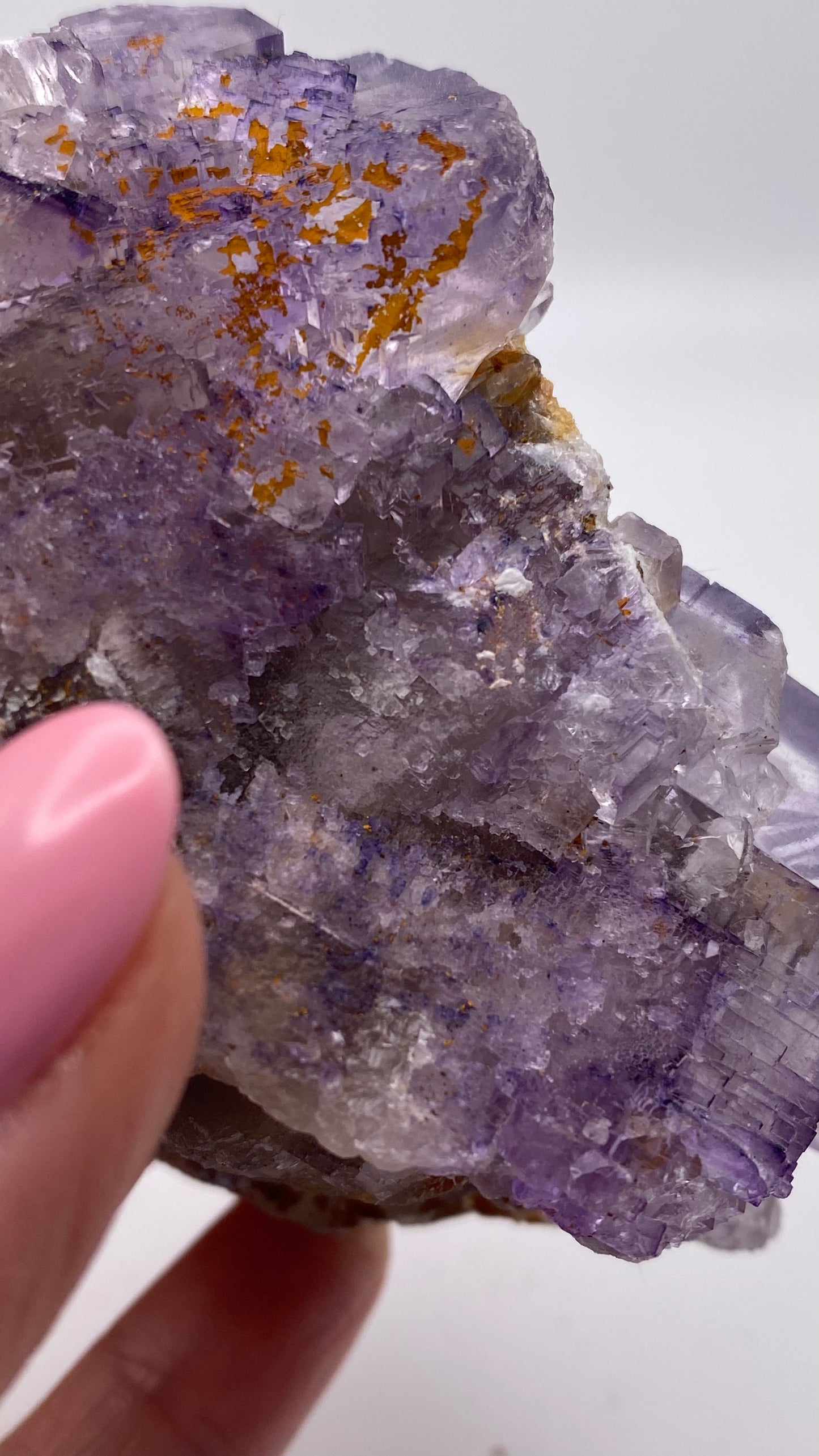 A close-up shot of a person's hand holding a Fluorite crystal from Lead Hill, Cave-In-Rock, Hardin County, Illinois, by The Crystalary. The person's nails are painted pink, and the crystal showcases intricate, translucent formations with patches of yellowish-brown inclusions. The background is a plain white.