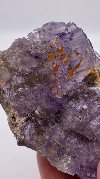 Close-up of a large, rough fluorite crystal held by a hand. The Fluorite- Lead Hill, Cave-In-Rock, Hardin County, Illinois, USA crystal from The Crystalary showcases a mix of vibrant purple and translucent hues with several patches of orange-brown mineral deposits scattered across its surface. The background is plain and out of focus, reminiscent of specimens from the renowned Cave-In-Rock Mining district in Hardin County.