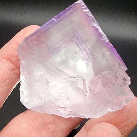 A person holding a translucent, slightly purple-tinted **Fluorite from Lead Hill, Cave-In-Rock Mining Sub-District, Hardin County, Illinois, USA** against a dark background. The crystal by **The Crystalary** has sharp edges and a triangular shape, with internal reflections highlighting its intricate structure.