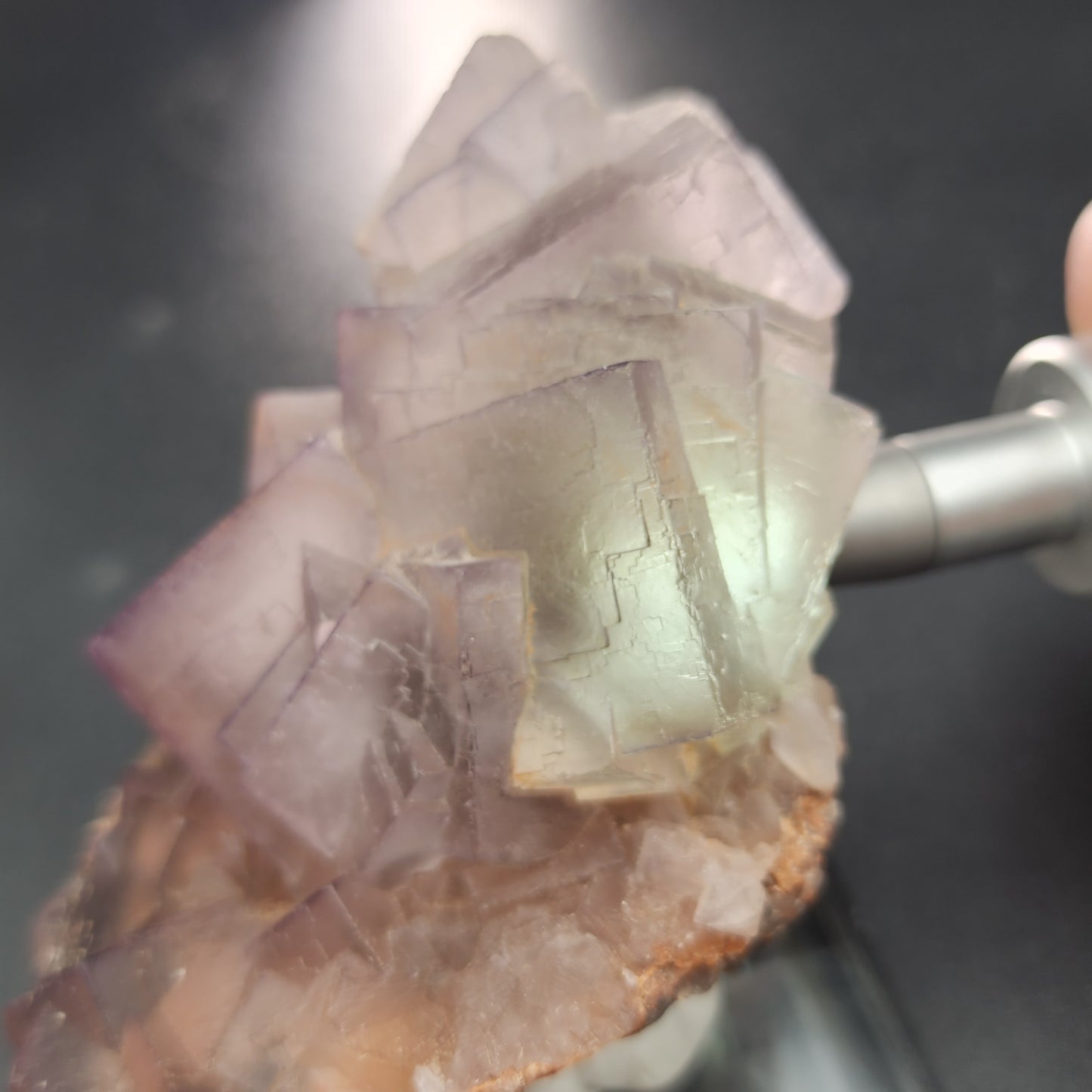 A close-up photo showcases a cluster of translucent, high-quality Fluorite crystals, exhibiting light purple and green hues. The intricate and delicate structure is perched on a beige, rough-textured base. Illumination from the right adds a soft glow to the colorful Fluorite crystals from The Crystalary's exquisite collection, sourced from Mekhtar, Loralai in Balochistan, Pakistan.