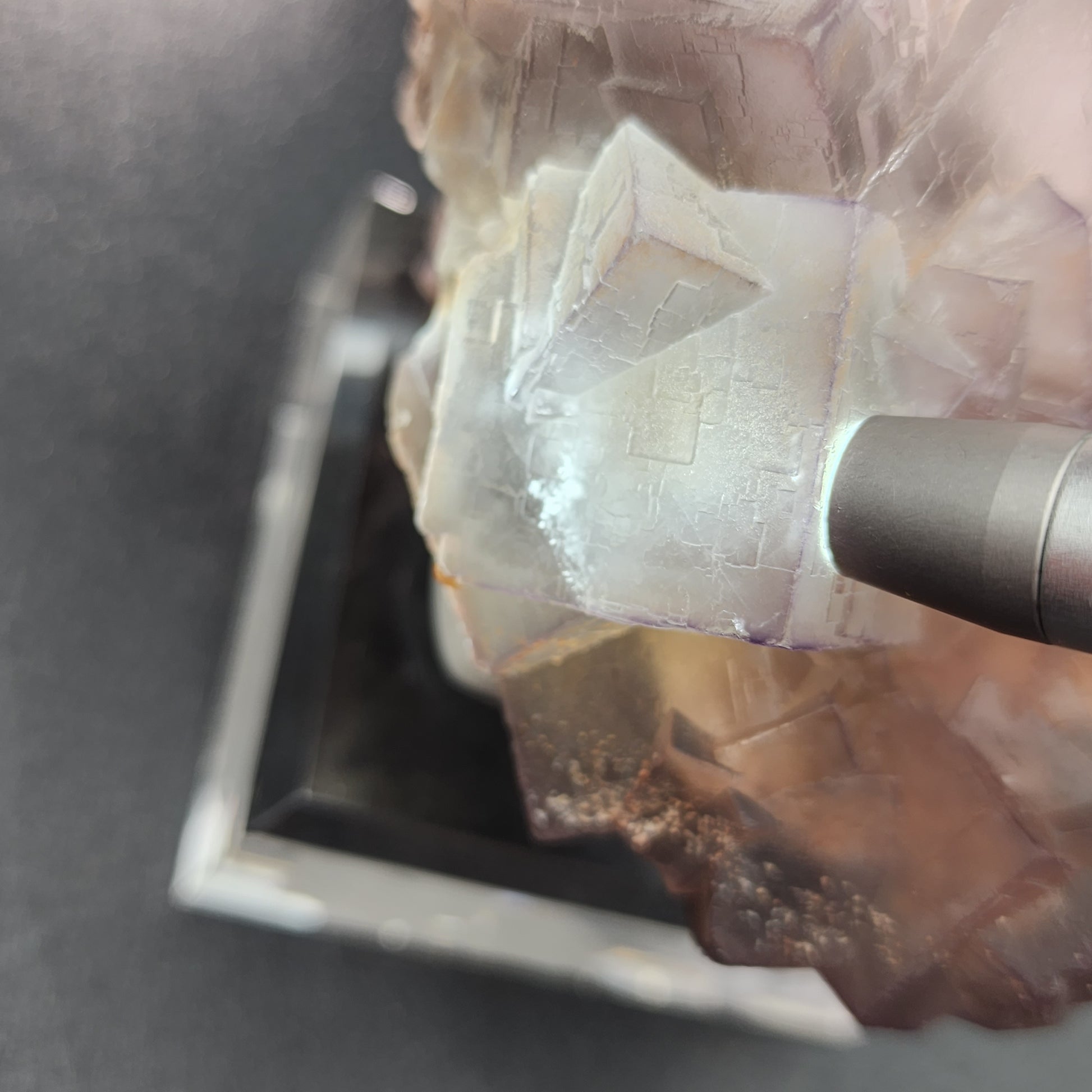 Close-up of a purple Fluorite, Mekhtar, Loralai, Balochistan, Pakistan mineral cluster by The Crystalary on a black reflective surface, with a small cylindrical object, likely a pen, pointing toward it. The minerals display a translucent, crystalline structure.