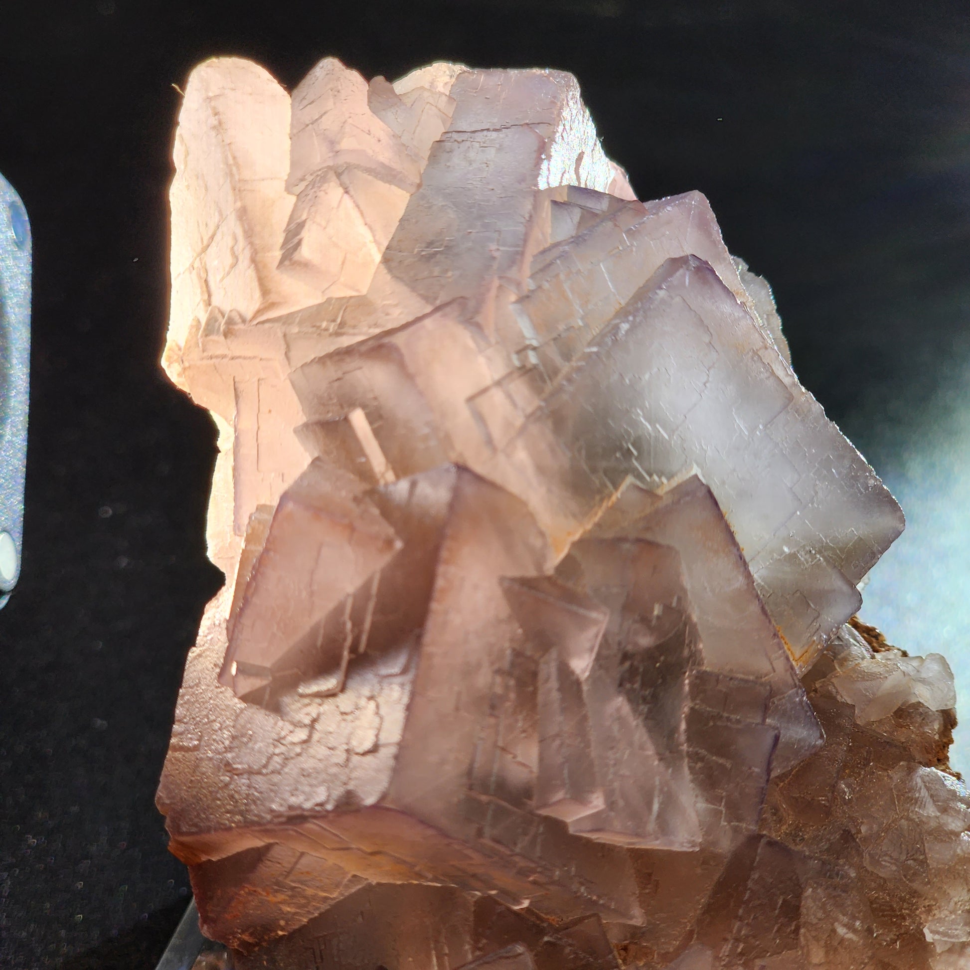 A close-up image of The Crystalary's fluorite from Mekhtar, Loralai, Balochistan, Pakistan, showcasing colorful crystals with interlocking cubic formations illuminated by a light source from the left side. This high-quality fluorite exhibits a light purple hue with some areas appearing almost clear and rests against a dark background.