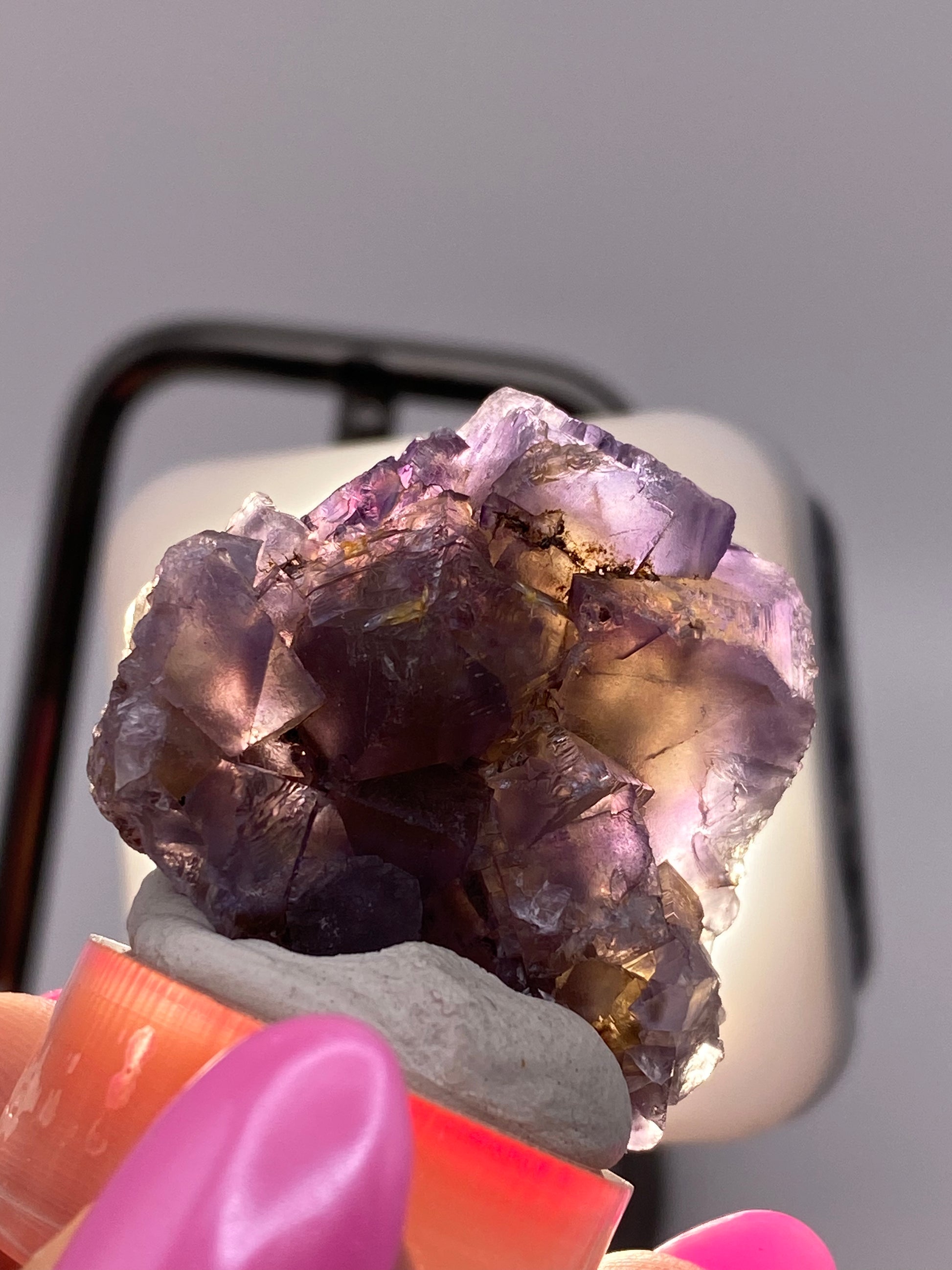 A hand with bright pink nails holds a cluster of purple cubic crystals against a blurred background. The crystals, likely Fluorite from Minerva Mine #1 in Hardin County, Illinois by The Crystalary, have a mix of translucent and opaque areas, including light violet partial cubes, and are mounted on a small clay base.