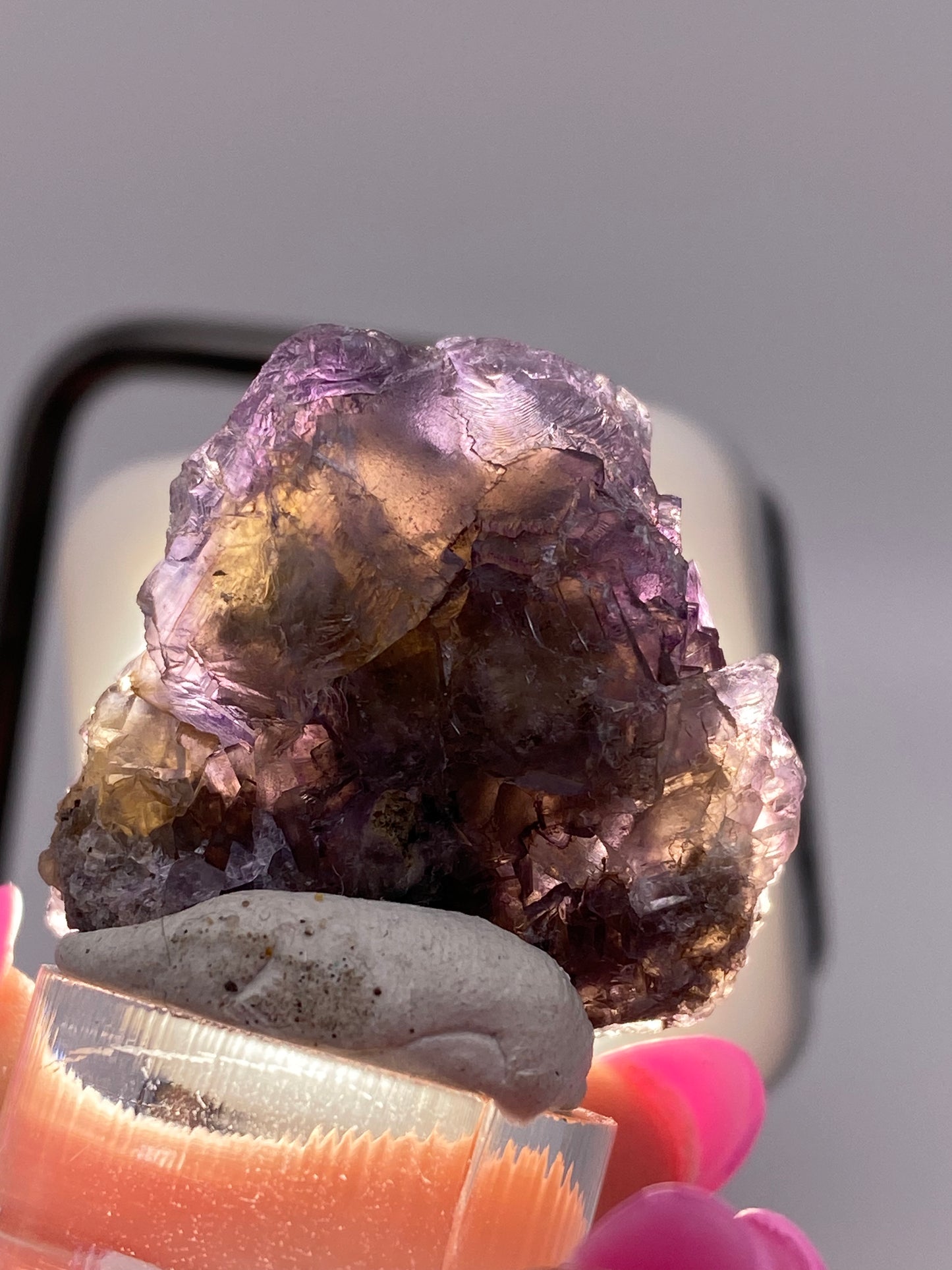 A close-up of a raw, irregularly shaped Fluorite from Minerva Mine #1 in Hardin County is held by a person with pink painted fingernails. The crystal, offered by The Crystalary, displays deep purple sections interspersed with lighter violet partial cubes and is showcased against a blurred, neutral background.