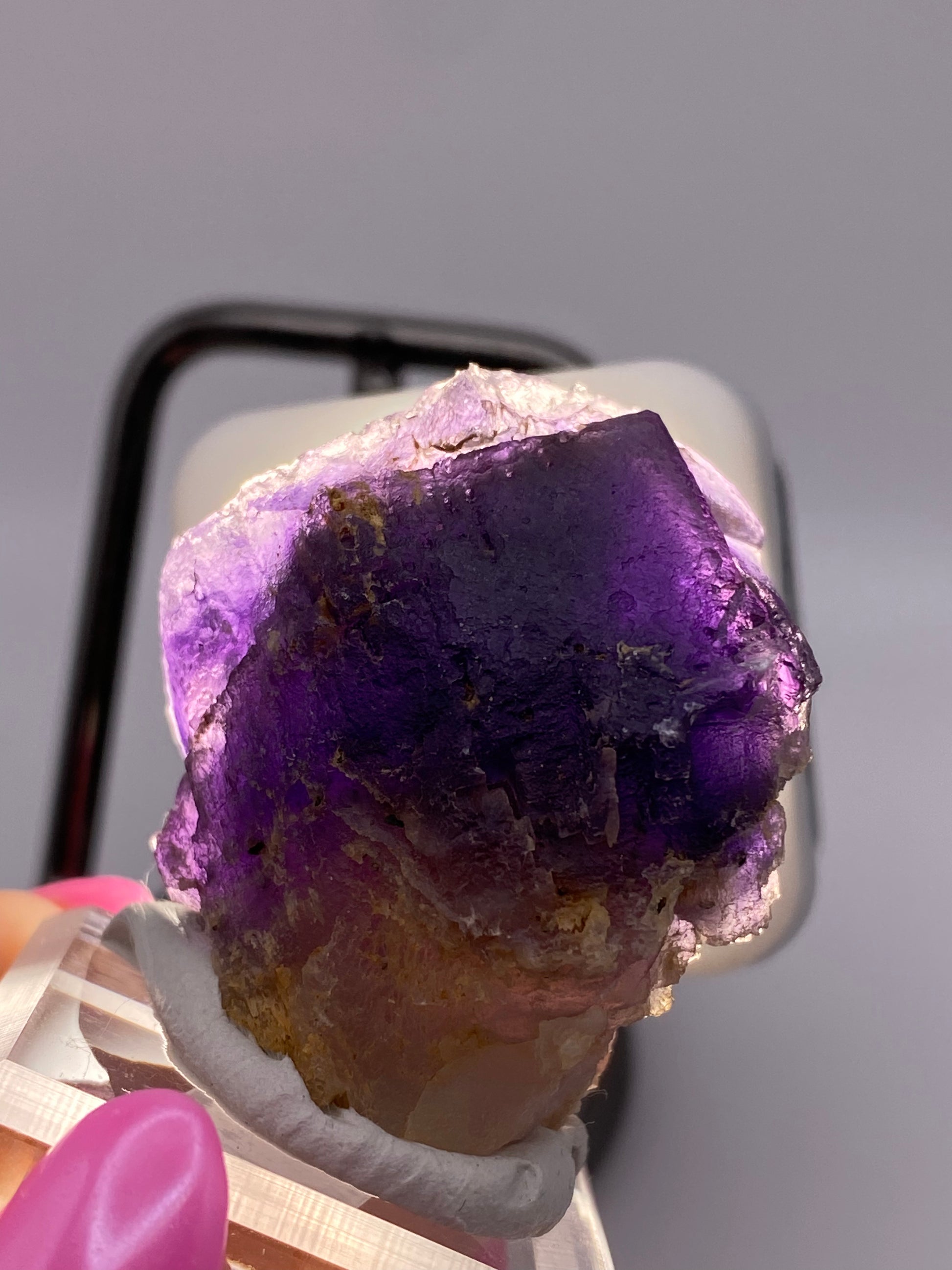 Close-up of a person's hand holding a Fluorite crystal from Minerva Mine #1, Hardin Co, Illinois by The Crystalary. The crystal displays violet to yellow hues in a partial cube form with white and brown inclusions. It is held against a neutral background and is illuminated to highlight its semi-translucent nature and detailed texture.