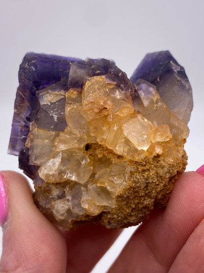 A hand with pink-painted nails holds a Fluorite specimen from Minerva Mine Number 1, Hardin County, Illinois. The specimen, available from The Crystalary, features translucent yellow and purple crystals with a rough, brown base. The crystal structure appears well-defined with a mix of sharp and slightly rounded edges, reflecting light like focal cubes.