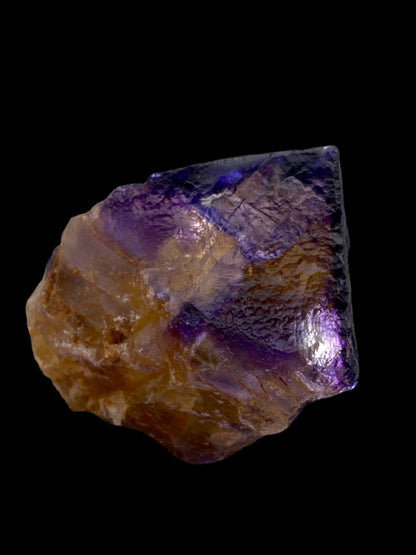 A close-up of a Fluorite specimen from Minerva Mine Number 1 in Hardin County, Illinois, marketed by The Crystalary, featuring deep violet partial cubes with a mix of purple and brown hues. The mineral displays a rough, faceted texture with sparkling surfaces set against a black background.