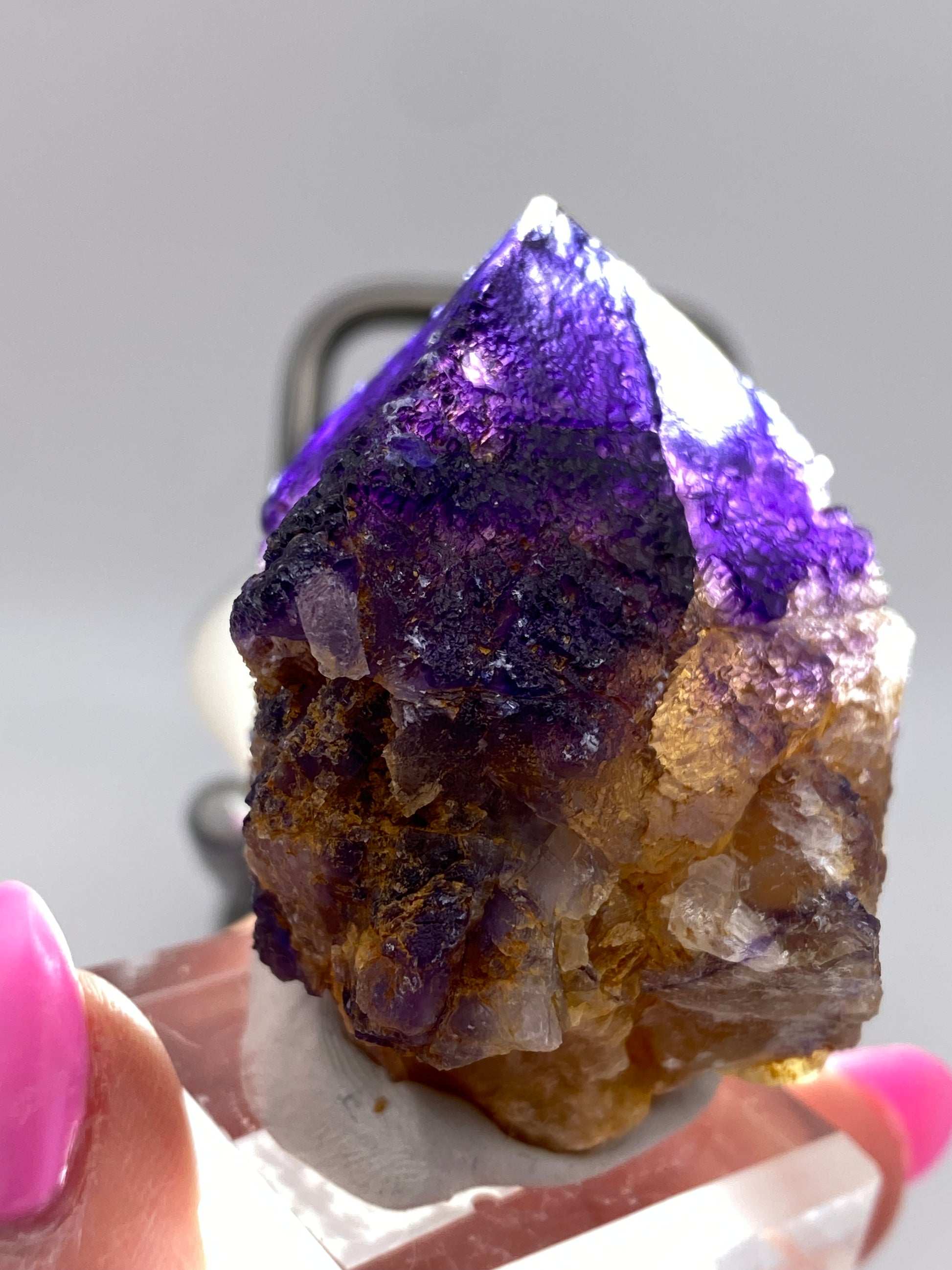 A close-up image captures a person's hand showcasing a Fluorite crystal from The Crystalary. With a blend of purple and light brown hues, this translucent Fluorite—sourced from Minerva Mine Number 1 in Hardin County, Illinois—displays its naturally formed, sharp edges and rough texture. The hand features pink nail polish, while the background remains blurred.