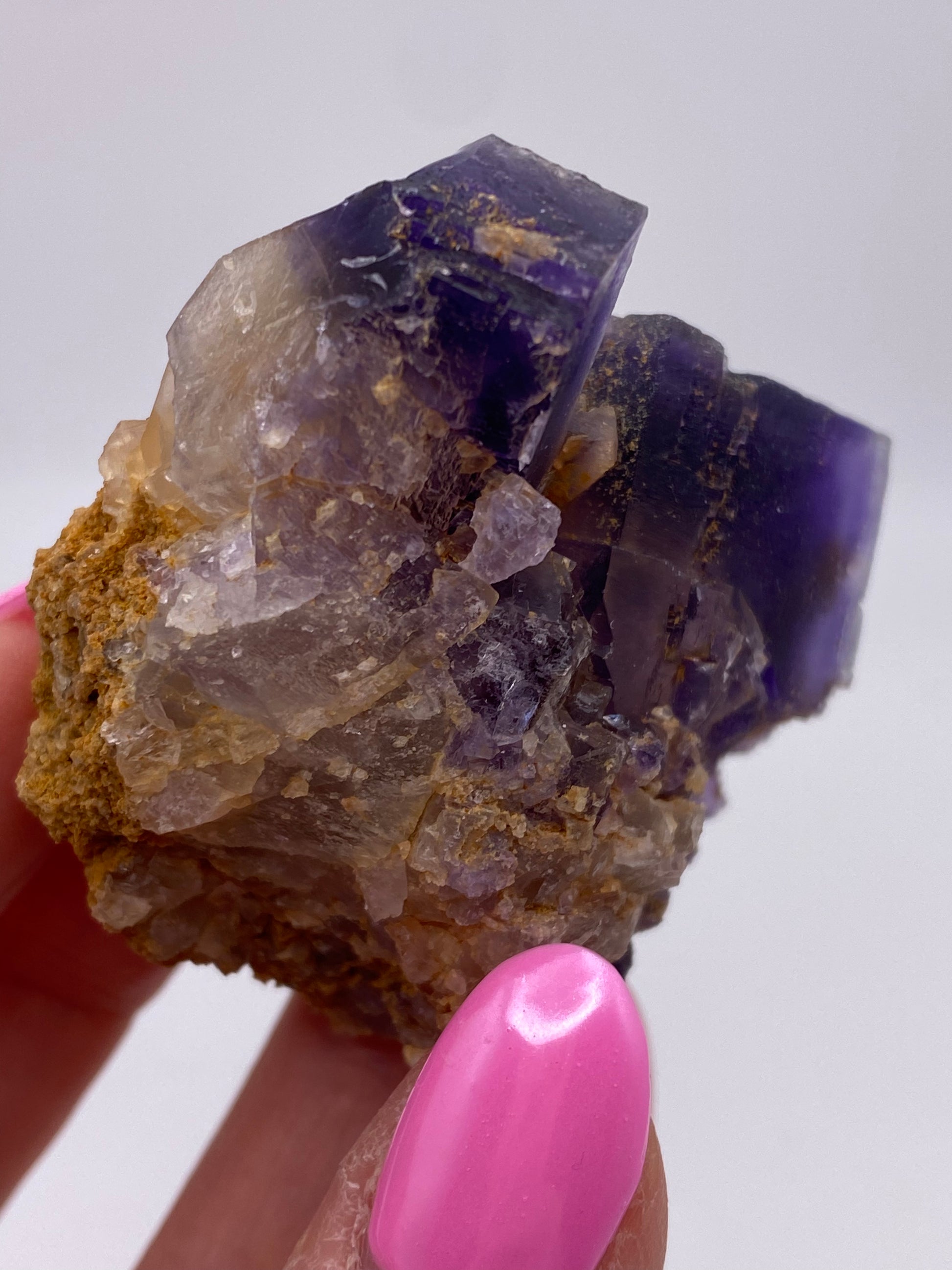 A close-up shot of a person holding a piece of raw, jagged Fluorite crystal from the Minerva Mine Number 1, Hardin County, Illinois by The Crystalary. The crystal features purple and translucent sections and is partially embedded in a tan, rocky matrix. The person's nails are painted bright pink. The background is white and out of focus.