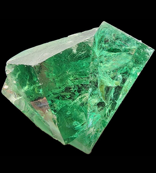 A close-up shot of The Crystalary's Fluorite Mini from the Diana Maria Mine in Frosterley, Weardale, Co. Durham, England. This vibrant green fluorite twin crystal exhibits a transparent, glass-like appearance with smooth faces and sharp edges. It showcases various internal fractures and inclusions, all set against a black background that highlights its remarkable clarity and color.