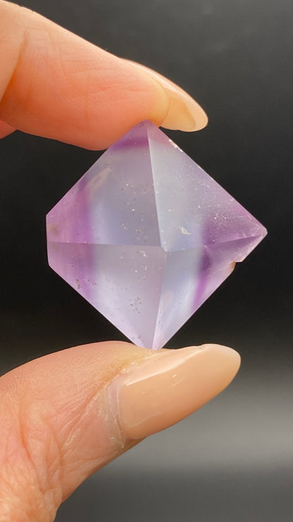 A hand with manicured nails holds The Crystalary's Fluorite Octahedron from Cave-in-Rock, Hardin Co, Illinois against a dark background. This purple, translucent crystal features a faceted geometric shape with slight internal imperfections and color variations.