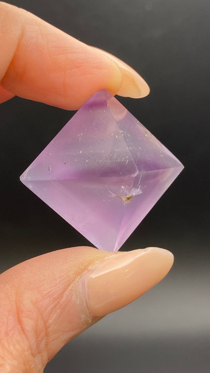 A close-up of a hand holding a translucent, lavender-colored, diamond-shaped crystal from The Crystalary against a dark background. The Fluorite Octahedron, sourced from Cave-in-Rock in Hardin Co, Illinois, has a small inclusion and is gently pinched between the thumb and index finger of a person with a polished manicure.