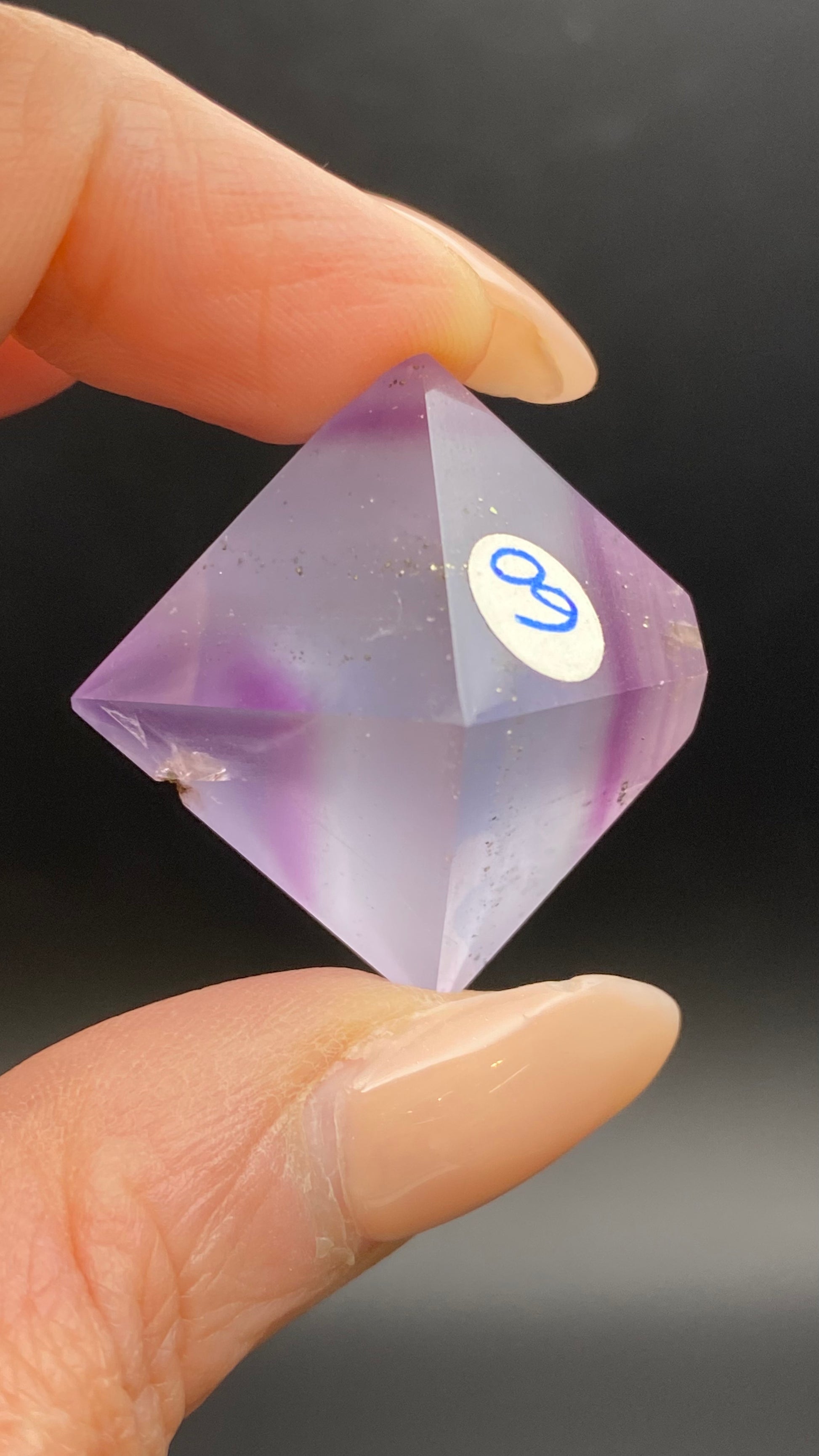 A hand with manicured nails holds a Fluorite Octahedron from Cave-in-Rock, Hardin Co, Illinois by The Crystalary, labeled "9" on its surface. This hand-polished gemstone is cut into a symmetrical, multi-faceted shape with semi-transparent, slightly cloudy areas. The background is dark and out of focus.