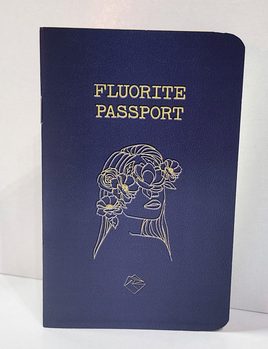 A dark blue "Fluorite Passport Book" by The Crystalary features gold lettering on the cover. An outline drawing of a woman's face adorned with flowers and hair, also in gold, graces the front. Perfect for logging your fluorite collection, it includes spaces for custom locality stickers and a small geometric logo at the bottom.