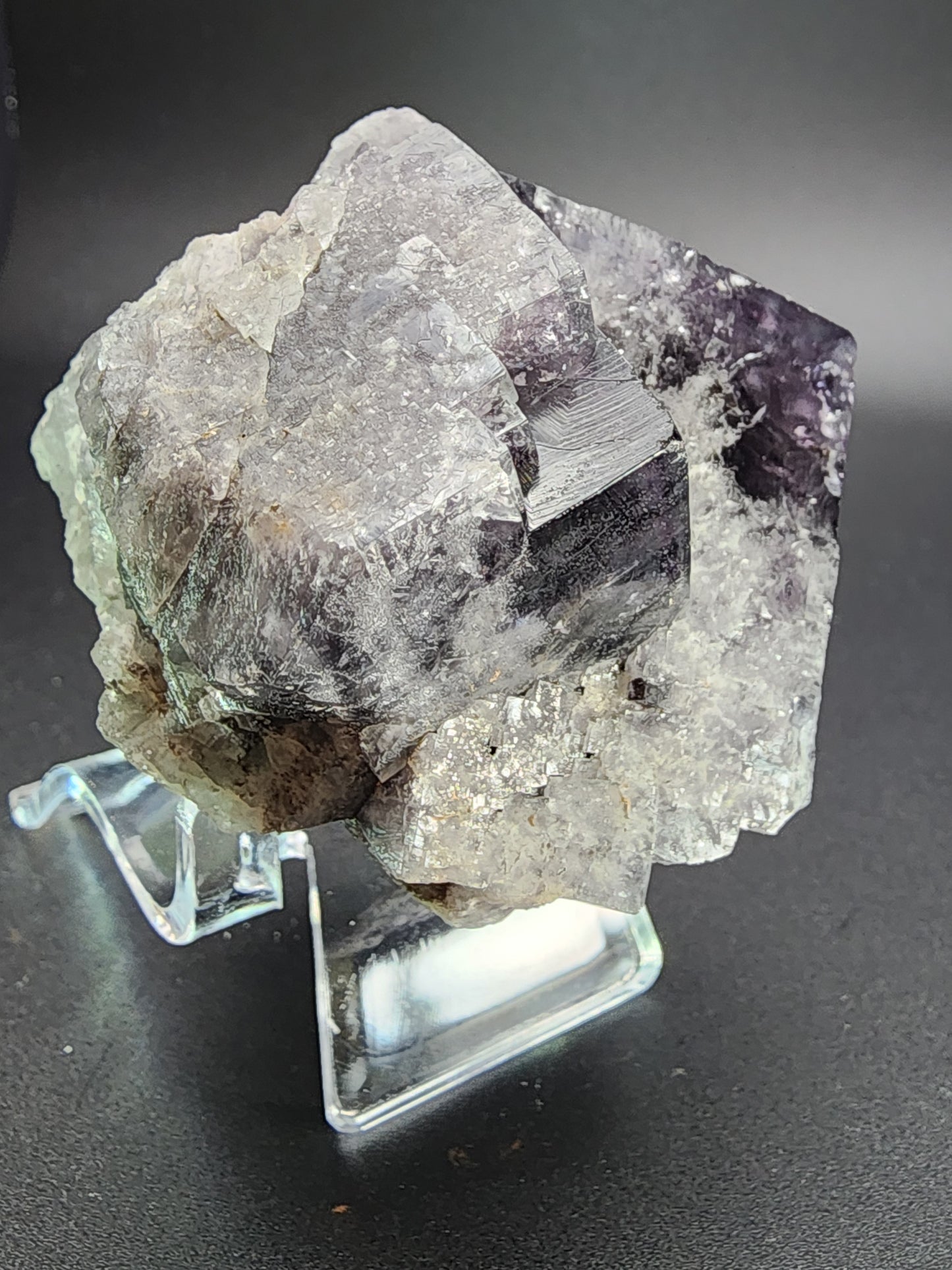 A close-up image of a large, rough fluorite crystal from The Crystalary's Purple Rain Pocket collection, sourced from the Fairy Hole Vein in the Lady Annabella Mine, Eastgate, Weardale, Co. Durham, England, showcases a combination of purple and clear hues. The crystal is displayed on a clear plastic stand against a dark background to highlight its natural textures and colors often seen in premium fluorite specimens.