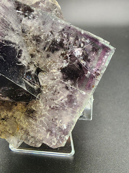 Close-up of a raw mineral specimen from The Crystalary, featuring a prominent rectangular, semi-transparent fluorite crystal with shades of purple, lavender, and gray. This Purple Rain Pocket fluorite from the Fairy Hole Vein in Lady Annabella Mine, Eastgate, Weardale, Co. Durham, England showcases jagged edges and intricate internal structures. It is displayed on a transparent stand against a dark background.