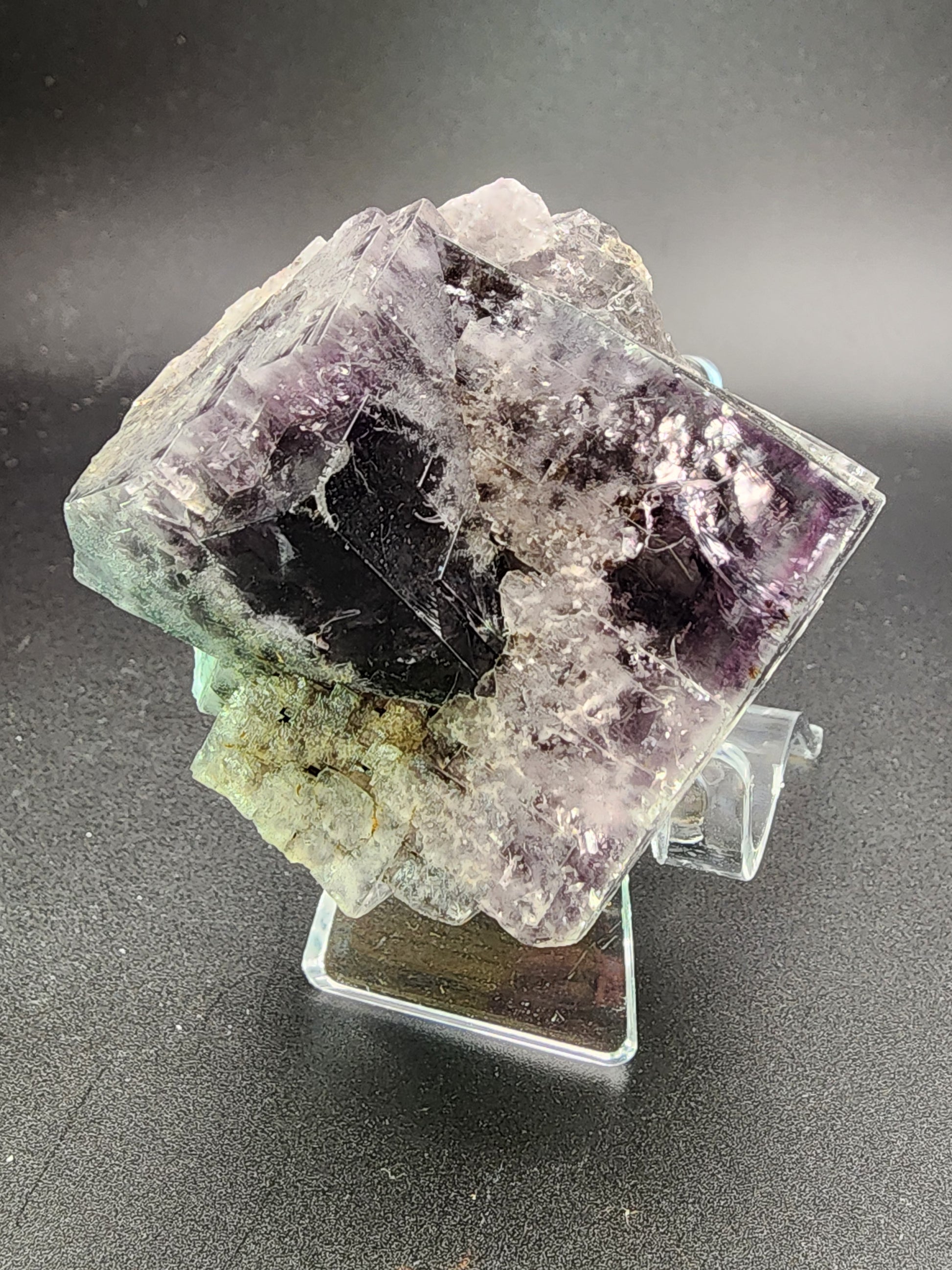 A close-up photograph of a transparent, cubic mineral specimen with a dark purple core from the Purple Rain Pocket, Fairy Hole Vein, Lady Annabella Mine in Eastgate, Weardale. The fluorite specimen from The Crystalary has a rough texture with visible cracks and a mix of light and dark patches. It is displayed on a clear plastic stand against a plain black background.