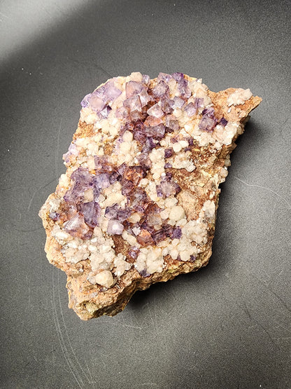 A close-up photograph of a rough mineral specimen featuring clusters of purple and white crystalline structures, identified as *Fluorite Quartz - Nonsense Pocket* from Crystal Peak, Park County, Colorado by The Crystalary, on a beige and brown rock matrix, displayed on a dark surface.