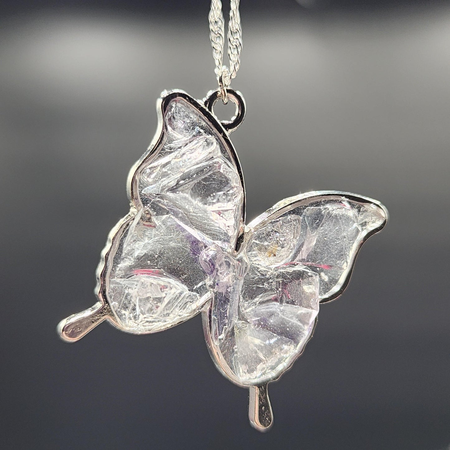 Close-up of the Fluorite Silver Butterfly Necklace, Cave In Rock from The Crystalary, a pendant shaped like a butterfly and made of clear, textured fluorite. The pendant is outlined in silver and hangs from a delicate silver chain against a dark, blurred background. This exquisite piece is an example of fine handmade jewelry.