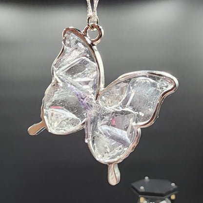 A close-up image of the Fluorite Silver Butterfly Necklace from The Crystalary. The pendant, featuring transparent, textured wings akin to ice or glass, is exquisitely crafted in silver. It dangles elegantly from a thin silver chain against a dark, plain background, highlighting the intricate beauty of this handmade jewelry piece from Cave In Rock.