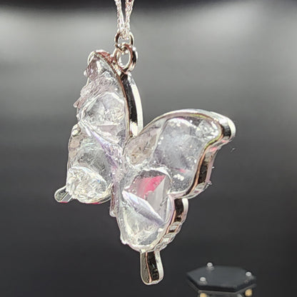 Close-up of the Fluorite Silver Butterfly Necklace, Cave In Rock by The Crystalary, featuring a clear butterfly-shaped pendant with silver edges hanging from a chain against a dark background. The necklace's textured, crystalline appearance evokes the beauty of Cave in Rock fluorite and reflects light, creating a delicate and elegant look.