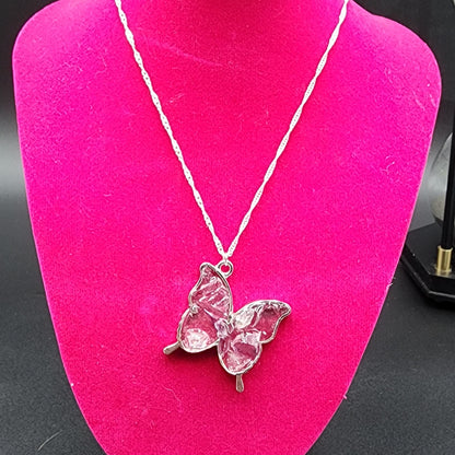 The Fluorite Silver Butterfly Necklace from The Crystalary is displayed on a bright pink mannequin. This stunning piece features a twisted chain and a pendant with pink, textured wings, embodying the elegance of handmade jewelry. The dark background beautifully highlights the delicate design of this Cave In Rock treasure.