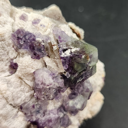 A detailed view of Fluorite, Smokey Quartz- Green Monster Pocket from The Crystalary showcases a mineral specimen featuring a stunning blend of purple and green fluorite crystals embedded in a white, rocky matrix from the Tarryall Mountains in Colorado. The irregularly shaped crystals feature translucent sections with varying hues, creating an intricate texture reminiscent of stained glass set against the contrasting background.