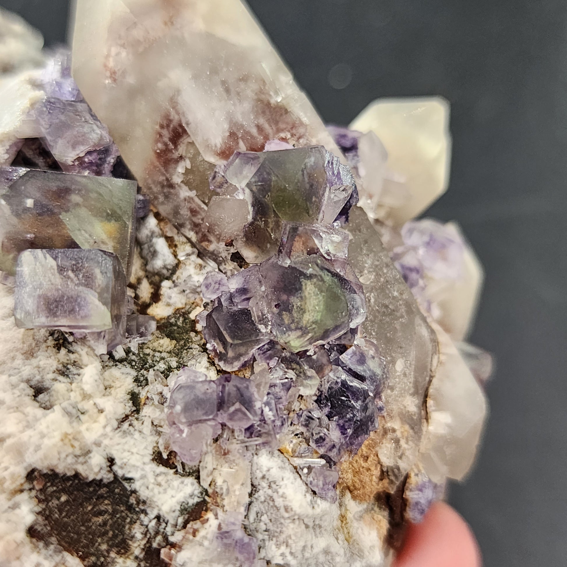 Close-up of a hand holding a mineral specimen, showcasing clear and purple cubic crystals of fluorite embedded in a larger, rough stone matrix. The background is blurred and dark, highlighting the intricate details and textures of the crystals and stone with a stained glass effect. The specimen features "Fluorite, Smokey Quartz- Green Monster Pocket" from the Tarryall Mountains in Colorado by The Crystalary.