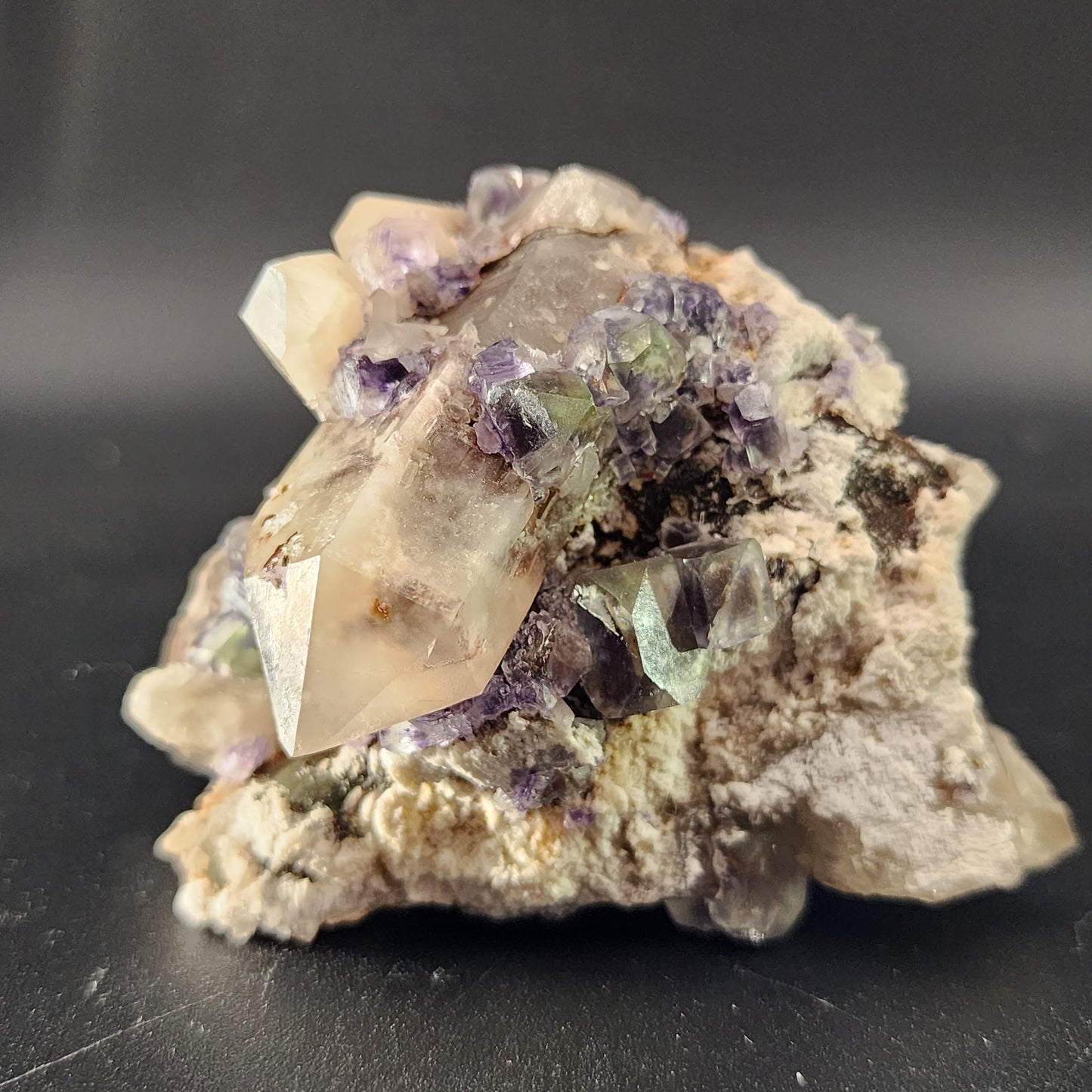 A close-up view of a "Fluorite, Smokey Quartz- Green Monster Pocket" from the Tarryall Mountains, Colorado by The Crystalary, featuring a cluster of quartz crystals adorned with purple and green fluorite octahedrons against a black background. The central smokey quartz crystal is large and partially transparent, surrounded by smaller crystals and mineral deposits that create a stunning stained glass effect.