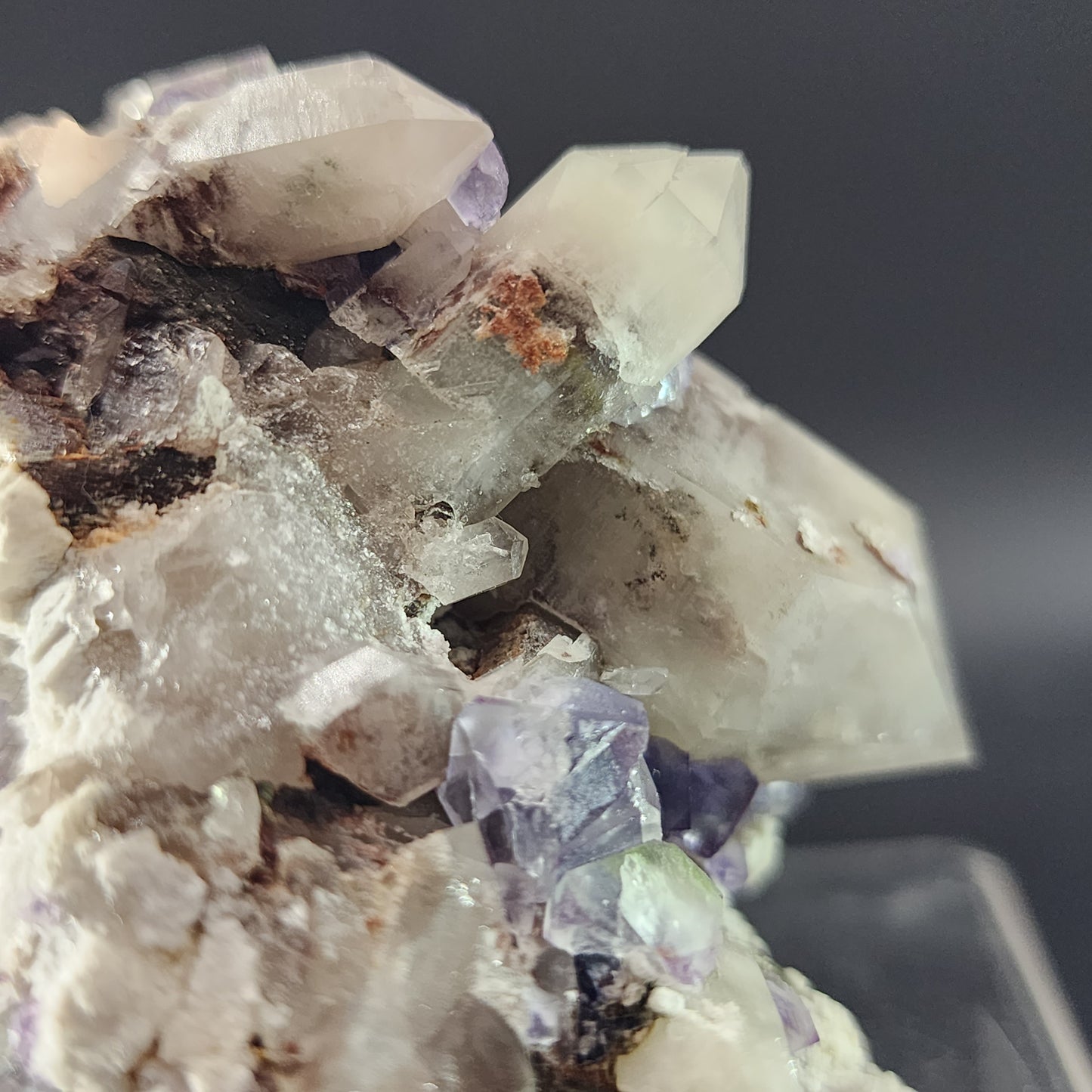 Close-up of a cluster of translucent fluorite and smokey quartz crystals embedded in a rocky matrix from the Green Monster Pocket in Tarryall Mountains, Colorado. The crystals range from white to light purple, with some showing sharp, pointed terminations. The dark background creates a striking contrast with the crystal formations, reminiscent of a stained glass effect. Available from The Crystalary.