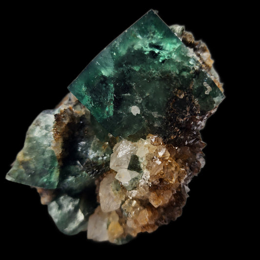 A detailed close-up of the Fluorite- Supernova Pocket crystal from Diana Maria, Co. Durham, England by The Crystalary. This green fluorite boasts rough, irregular surfaces and multiple translucent facets. It is set amidst a cluster of white and brownish mineral formations against a black background, enhanced by intertwined quartz crystals that add complexity to the beautiful composition.