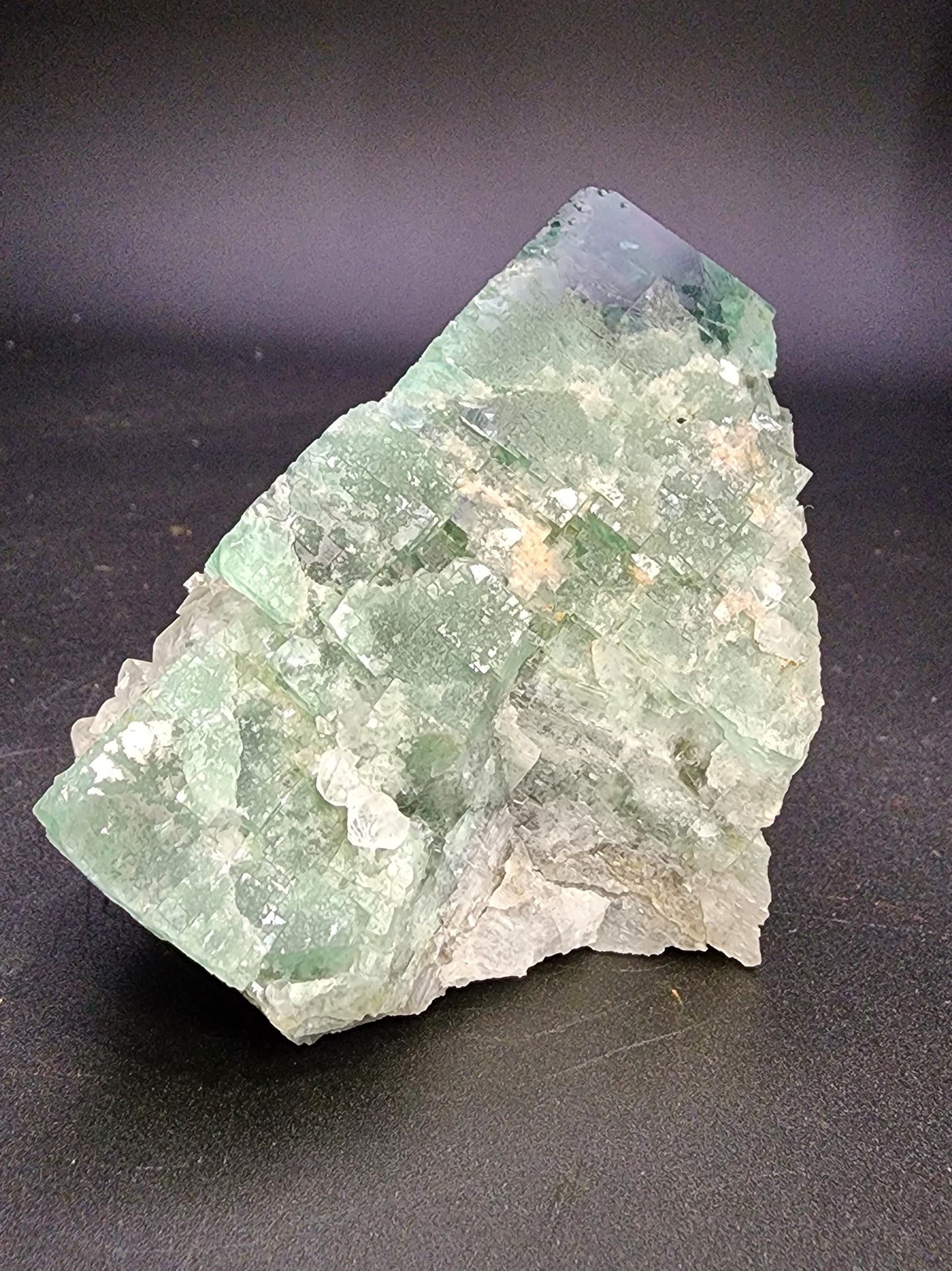 A large, rough specimen of Fluorite-Supernova Pocket from the Diana Marie Mine in Weardale, Co. Durham, England is displayed on a dark background. This crystal from The Crystalary features a pale green color with clear areas, jagged and uneven surfaces, numerous small facets, and a cloudy appearance in some regions.