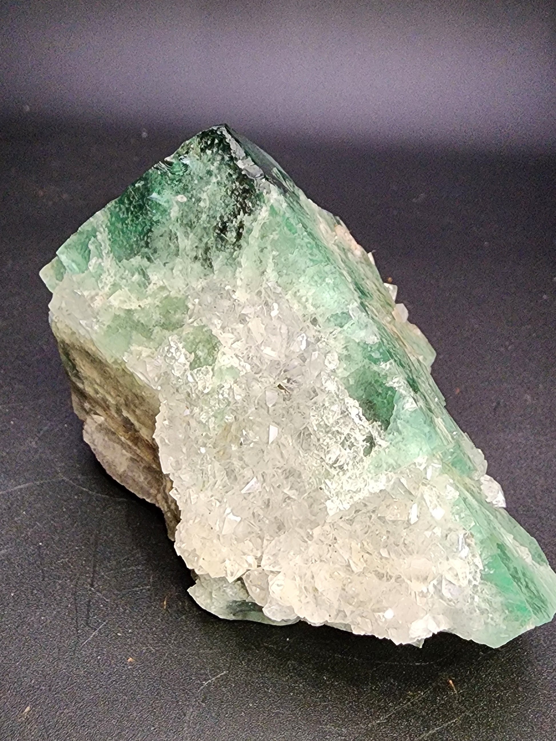 Close-up of a mineral specimen featuring a large chunk of green fluorite crystals with white crystalline formations from the Diana Marie Mine in Weardale, Co. Durham, England. The green fluorite has a translucent to opaque appearance, while the white crystals are more transparent and sparkly. This exquisite piece by The Crystalary, named Fluorite- Supernova Pocket, is set against a plain, dark background.