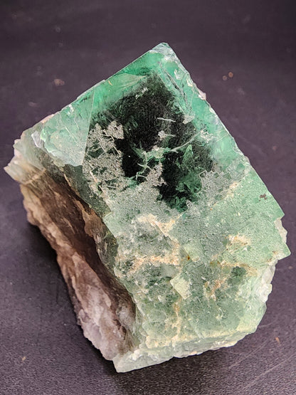 Close-up of a raw green fluorite crystal with hexagonal shape, offered by The Crystalary. This "Fluorite- Supernova Pocket" from the Diana Marie Mine in Weardale, Co. Durham, England, presents a mix of dark and light green hues accented by white and beige mineral deposits. The edges are rough, and the surface is partially translucent with visible crystalline formations inside that resemble those found in supernova pockets.