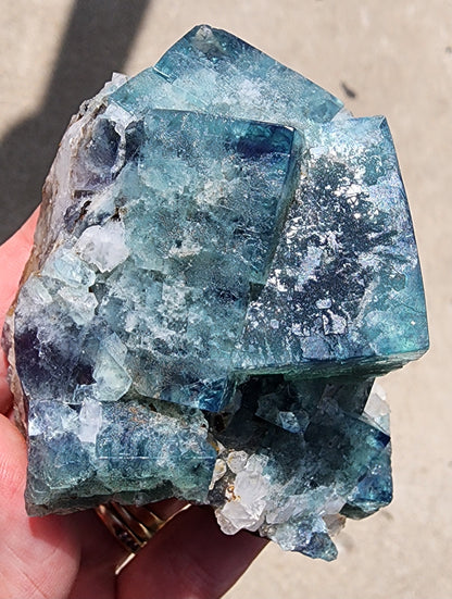 A hand is holding a Supernova Pocket Fluorite from The Crystalary, mined at the Diana Marie Mine in Weardale, Co. Durham, England. The large, rugged crystal formation exhibits stunning shades of blue with hints of white and possibly includes quartz crystals within its rough texture. Various fractures and reflections catch the sunlight beautifully, while the out-of-focus background highlights the intricate details of this exquisite piece.