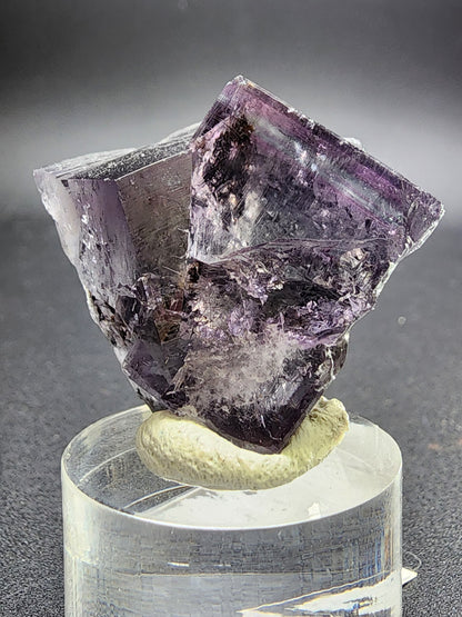 A detailed photograph of the Fluorite Twin, sourced from the renowned Purple Rain Pocket in Lady Annabella Mine, Eastgate, Weardale, Co. Durham, England, UK, is displayed on a clear pedestal. This translucent cubic crystal by The Crystalary features visible inclusions and natural imperfections that reflect light beautifully to showcase its intricate internal structure and interpenetrant twins.