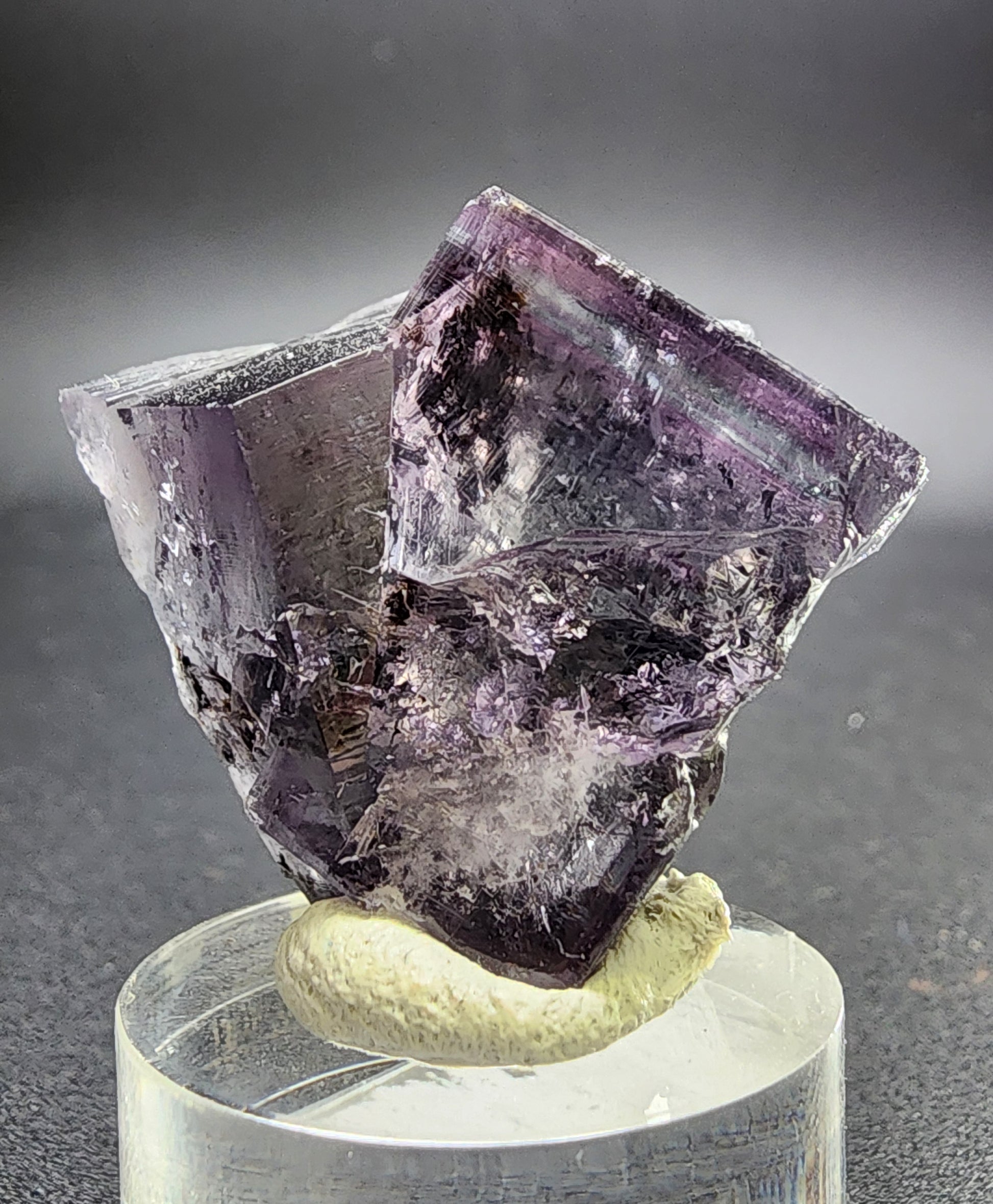 A close-up photo of a Fluorite Twin crystal specimen, sourced from the renowned Purple Rain Pocket in Fairy Hole Vein, Lady Annabella Mine, Eastgate, Weardale, Co. Durham, England (UK). Displayed on a clear stand and offered by The Crystalary, this purple fluorite crystal features visible facets and intricate internal patterns that highlight its transparent and translucent qualities. The blurred background emphasizes the detailed texture of the fluorite.