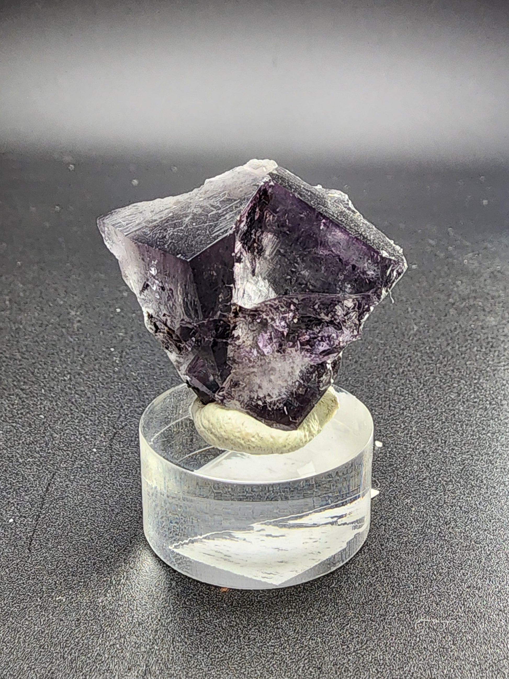 A Fluorite Twin crystal from the Purple Rain Pocket in the Fairy Hole Vein of Lady Annabella Mine is perched on a clear round display stand against a neutral background. This exquisite mineral specimen, offered by The Crystalary, features dark purple hues with sharp geometric shapes and a muted shine, making it one of the finest fluorites from Weardale, Co. Durham, England.