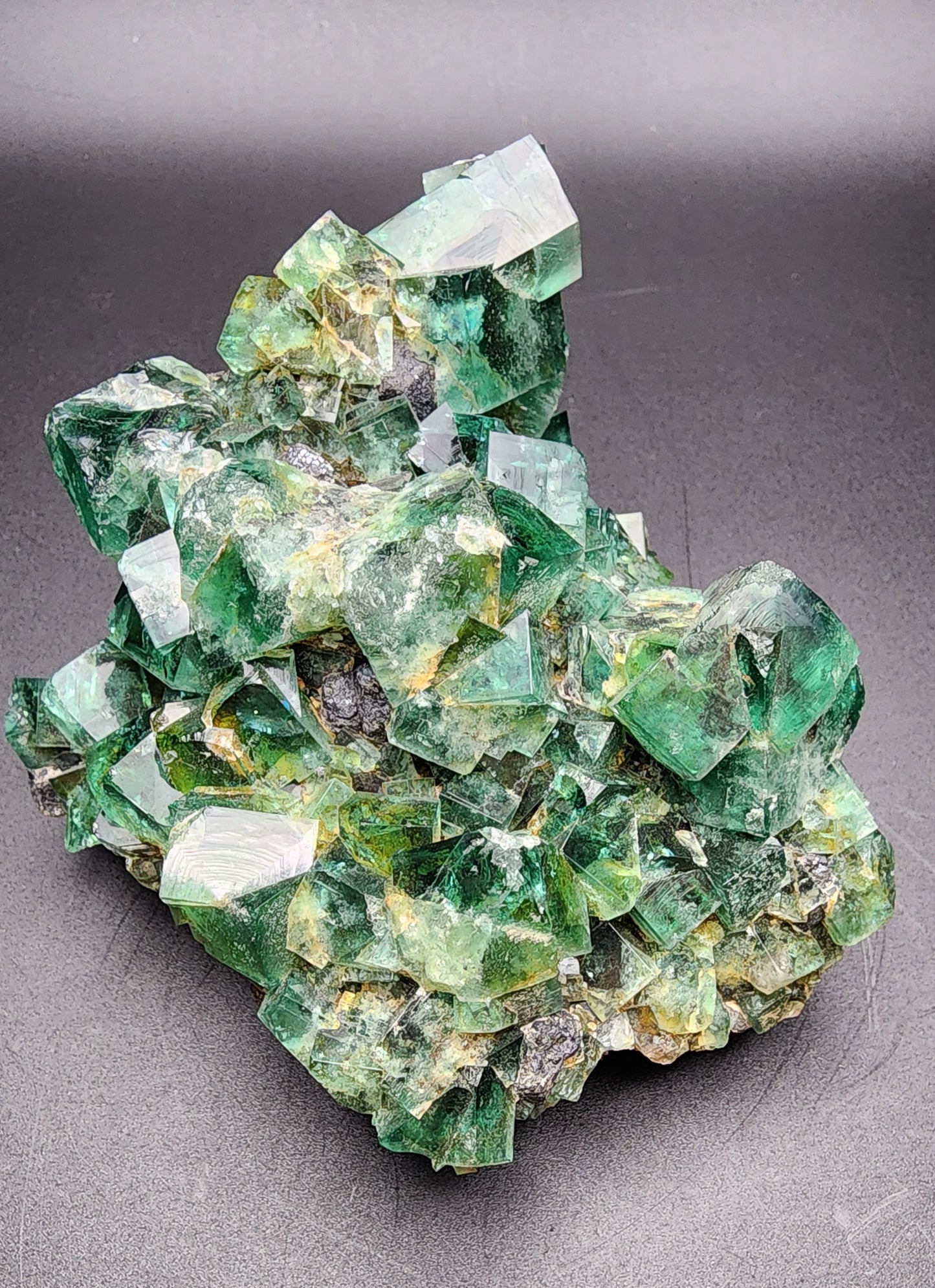 A stunning cluster of green fluorite crystals and a few galena crystals from The Crystalary's Fluorite with Galena (Heavy Metal Pocket) collection is showcased against a dark background. Emerging from the Heavy Metal Pocket in the Diana Marie Mine, Weardale, County Durham, England, these crystals vary in size and shape, exhibiting transparent to translucent qualities with intricate and sharp edges.